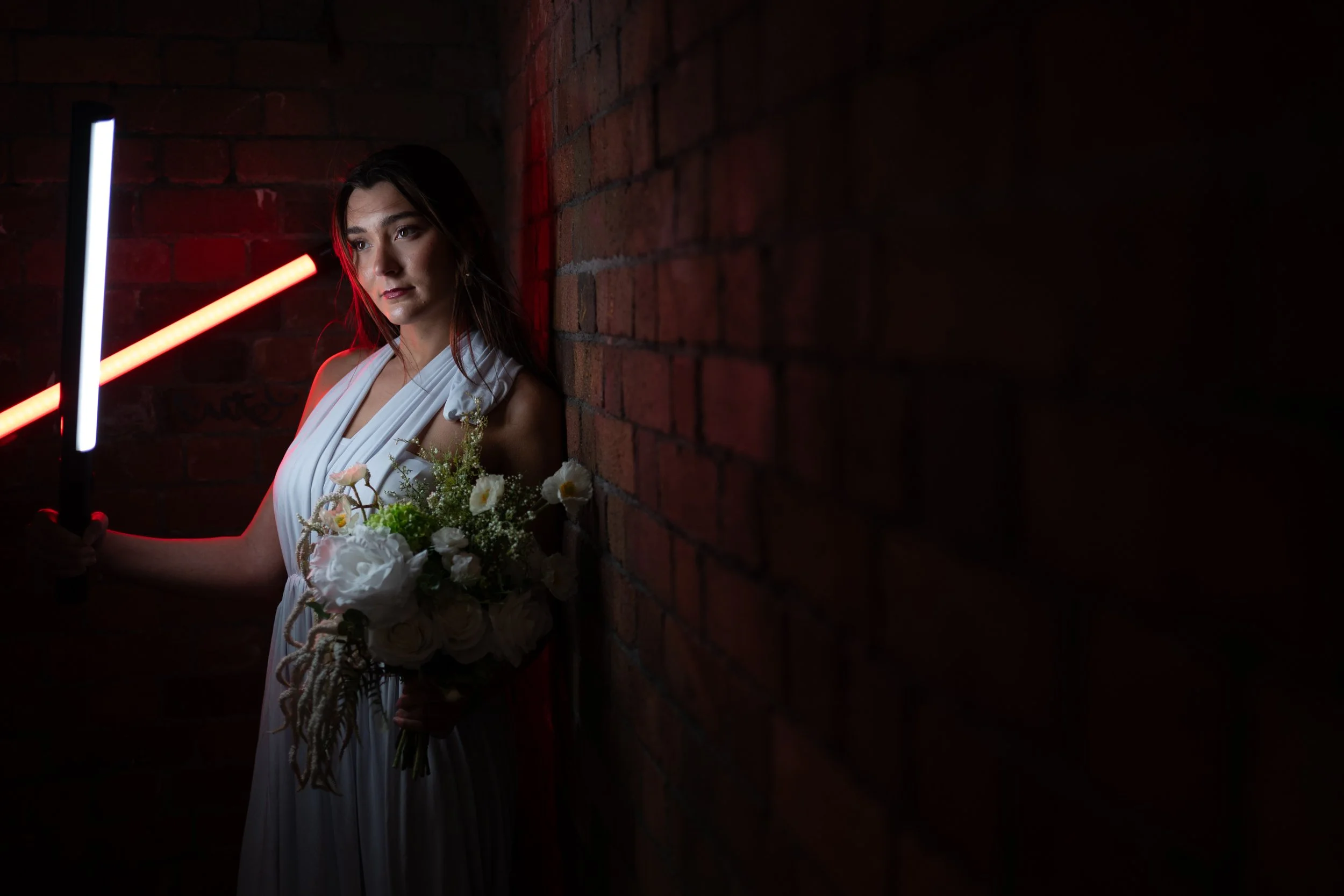 A woman in a white dress holding a bouquet of flowers, standing against a brick wall in a dimly lit setting with red and white neon lights.