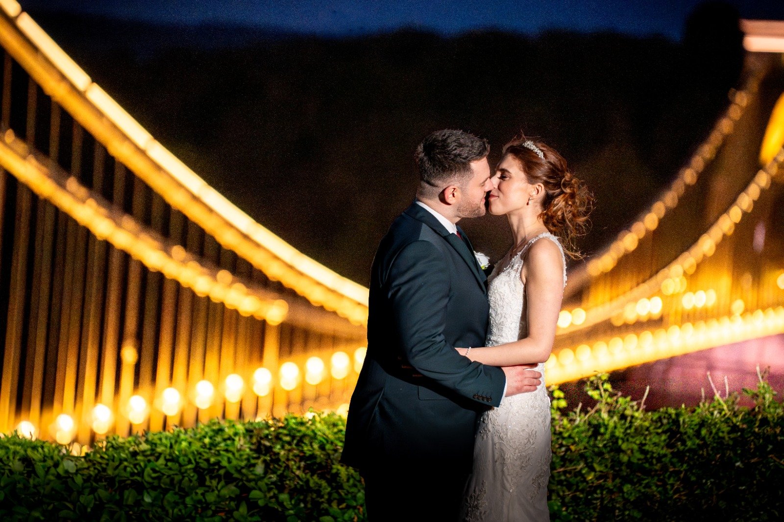 A bride and groom in wedding attire sharing a kiss at night on a bridge with illuminated lights in the background.