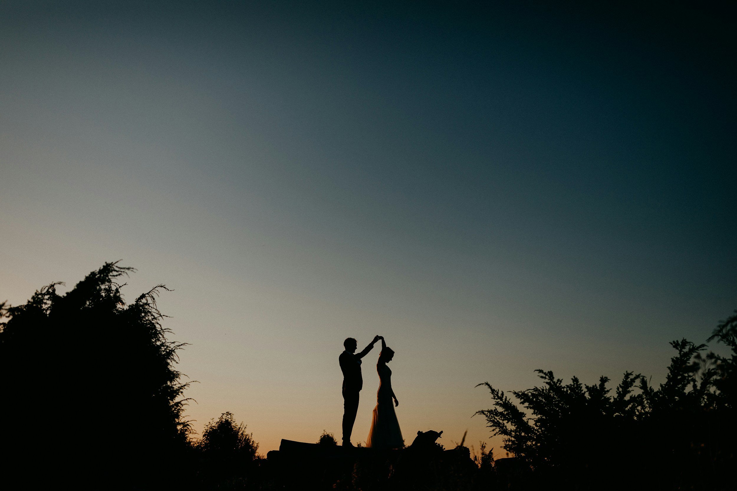 Silhouette of a couple during their wedding at sunset, with the groom placing a ring on the bride's finger, surrounded by trees against a colorful evening sky.