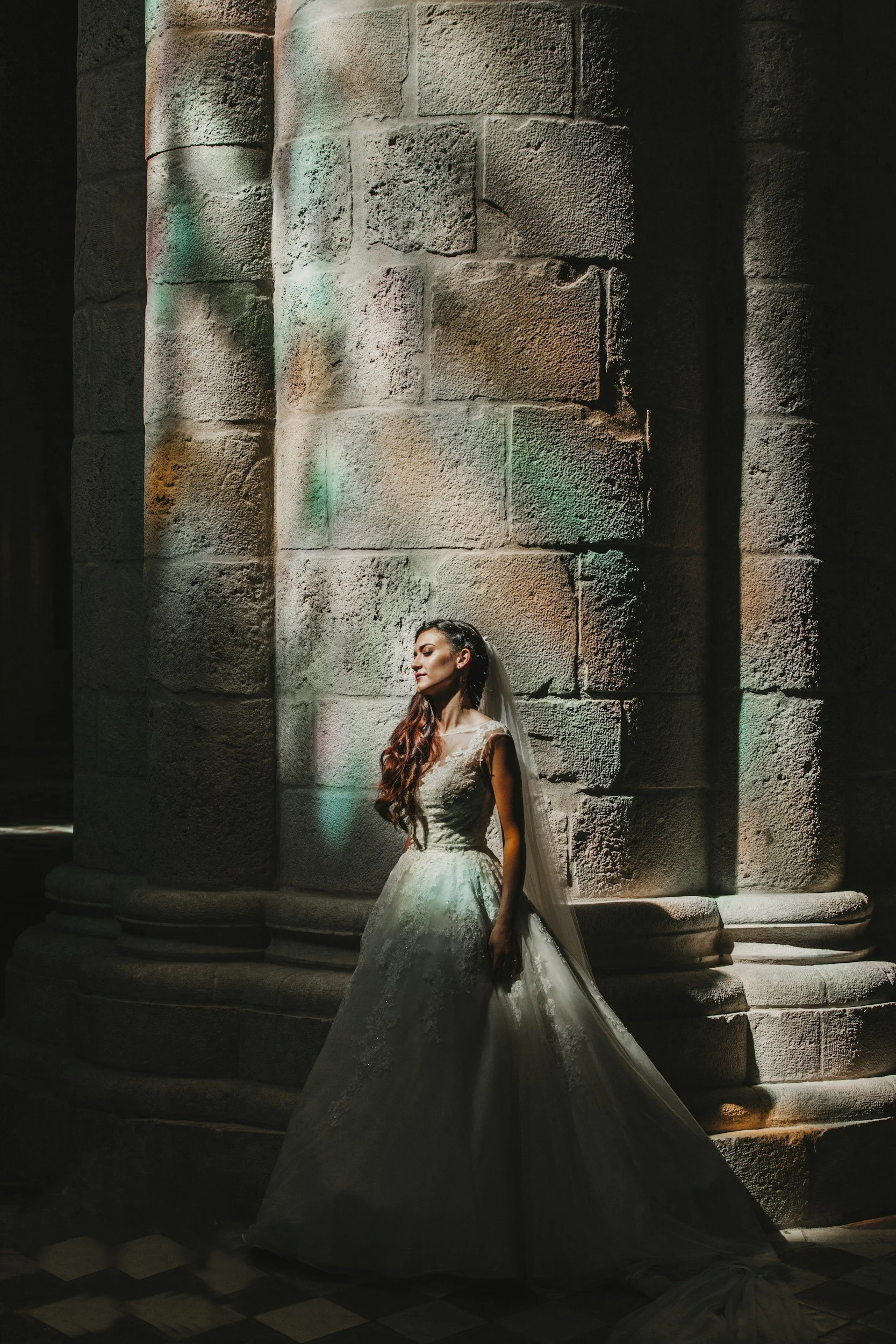 Bride in a white wedding dress and veil standing against a large stone column with colorful lighting reflections.