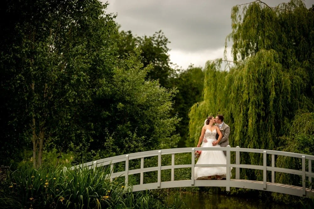 A bride and groom standing together on a small white bridge outdoors, surrounded by lush green trees and bushes. The couple is sharing a kiss.