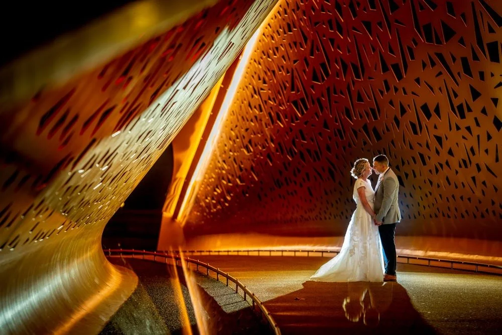 A bride and groom holding hands and looking at each other in front of modern, abstract architecture at night.
