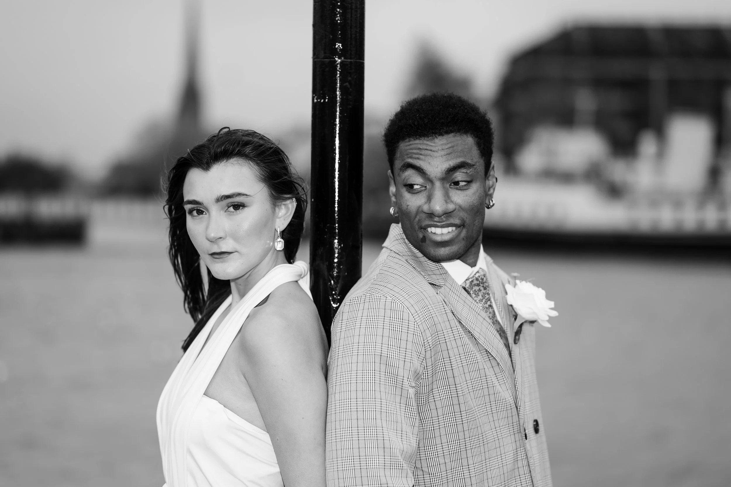 A black and white photo of a woman and a man standing back to back outdoors, separated by a metal pole, with a blurry cityscape background.