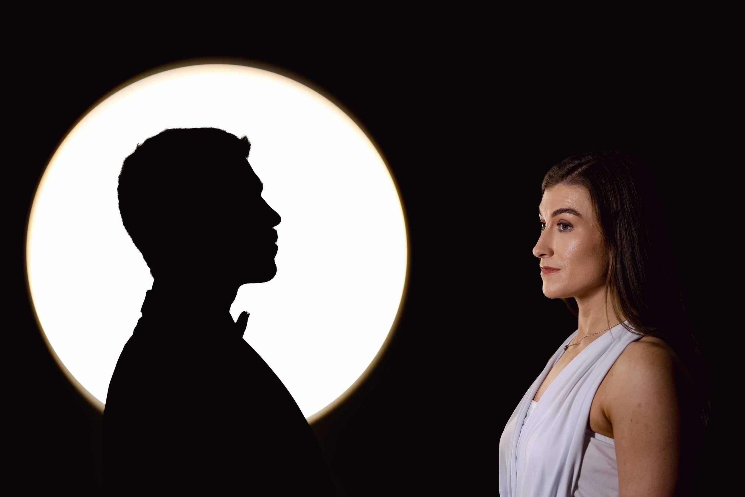 Profile of a woman in a white top with dark hair, facing a silhouette of a man in a tuxedo, with a bright circular light behind the silhouette, against a black background.