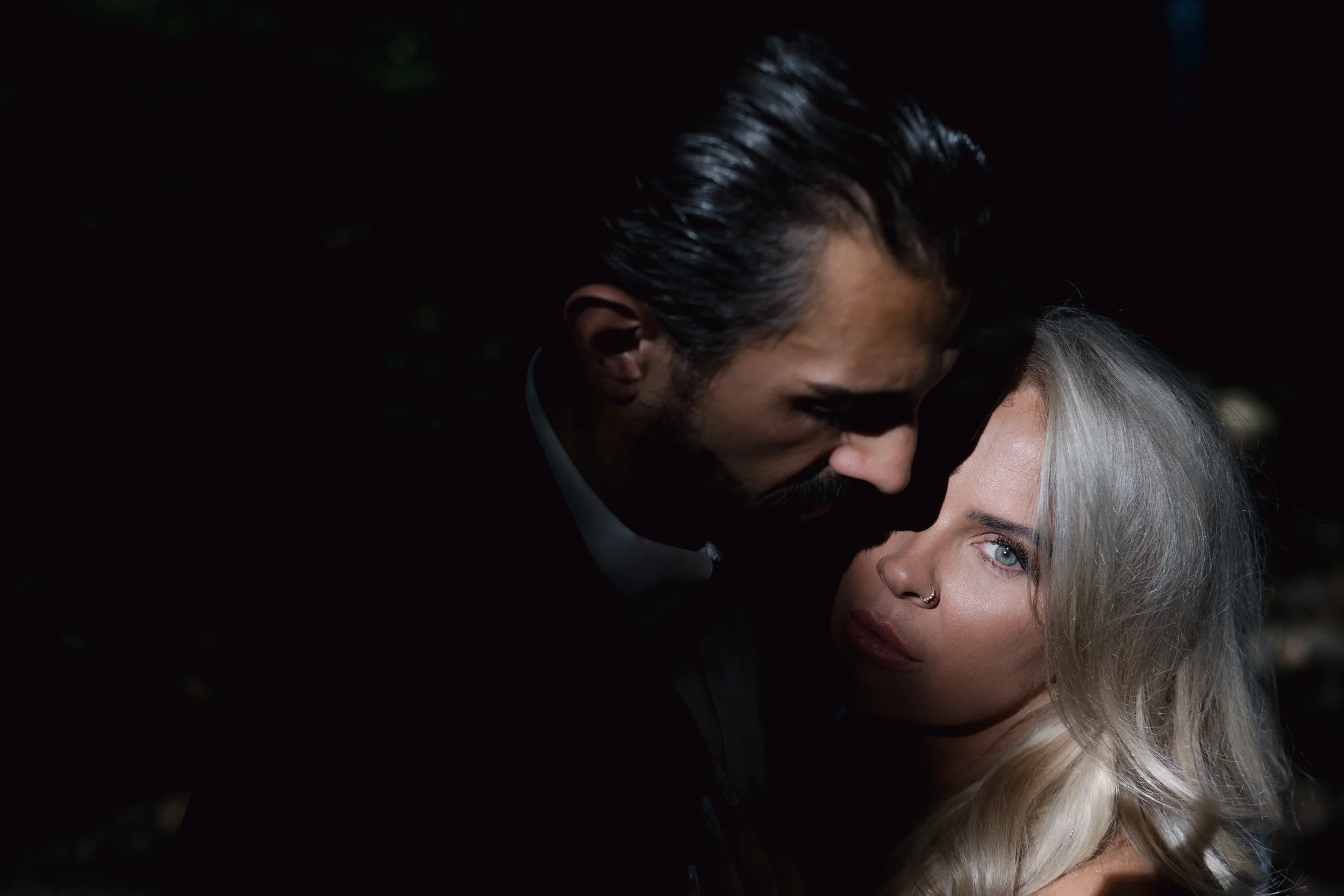 A man with dark hair and facial hair is leaning in to kiss a woman with light blonde hair and a nose piercing. The background is dark.