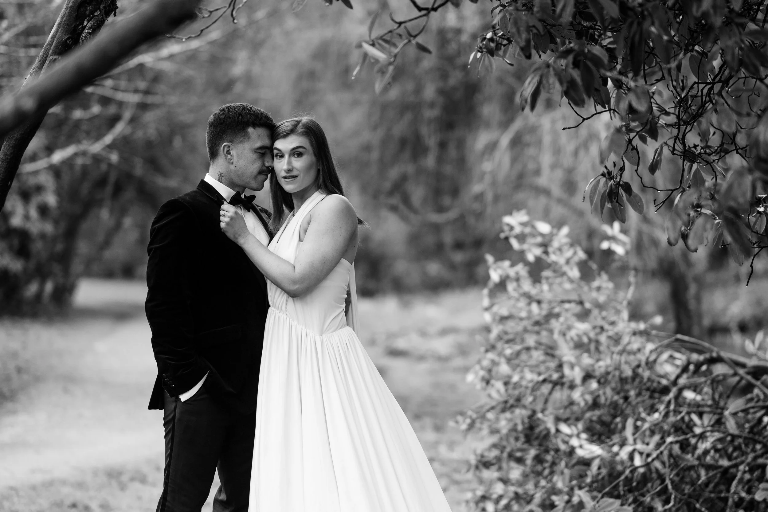 A black and white photo of a couple in formal attire, standing closely outdoors under trees, with the woman holding the man's lapel and both looking at the camera.