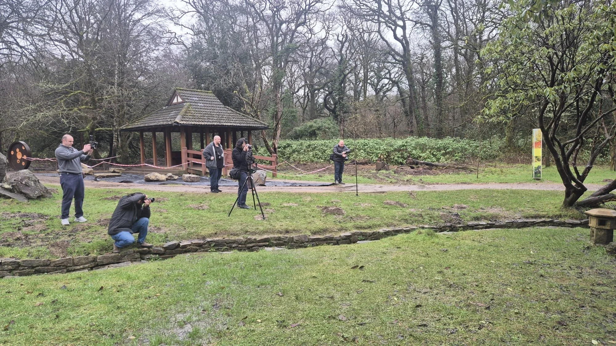 Group of photographers and videographers setting up equipment in a park on a rainy day, with a small gazebo and trees in the background.