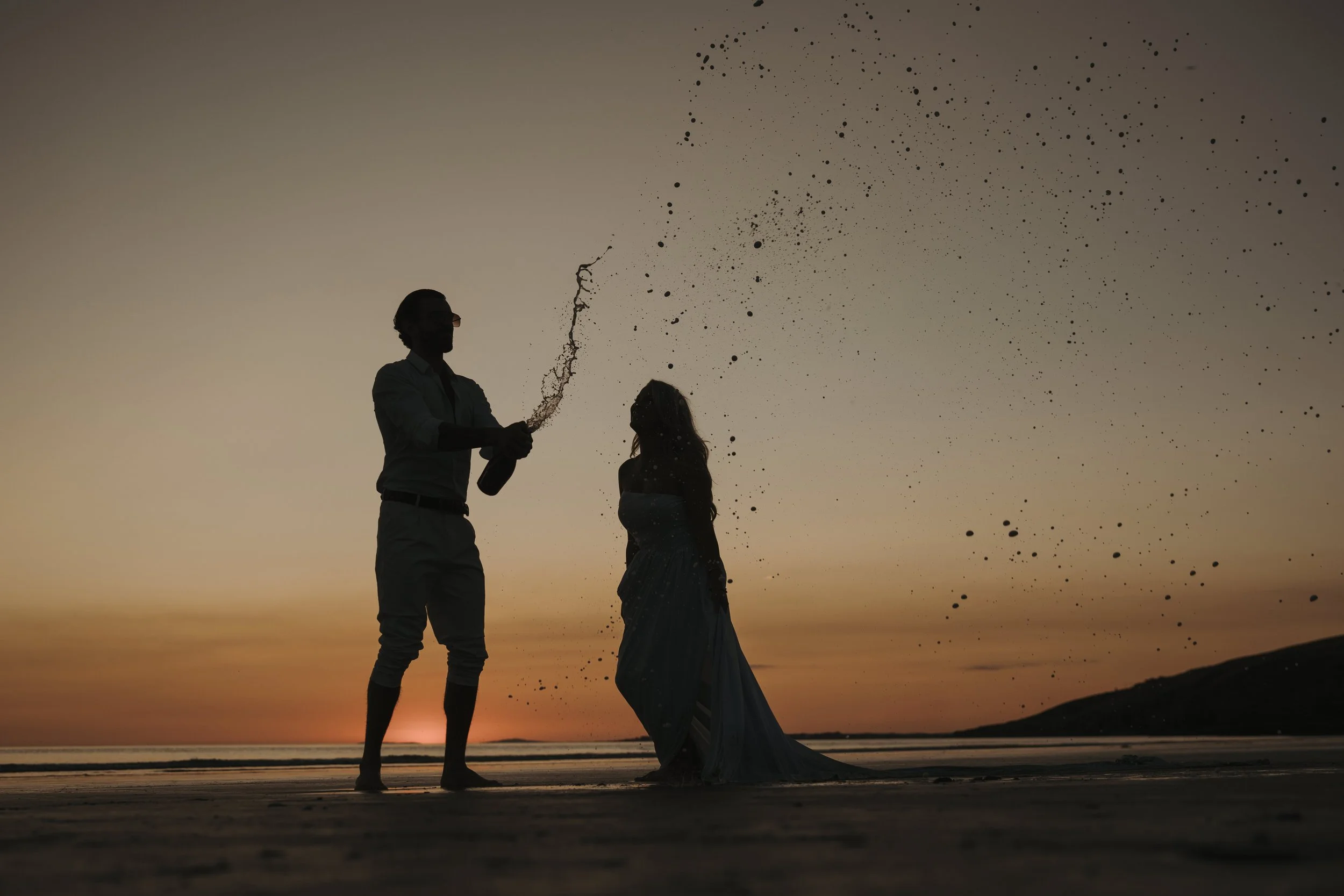 Silhouettes of a man and a woman on a beach at sunset, with the man holding a bottle and spray paint, and the woman wearing a dress, with a dark, starry sky in the background.