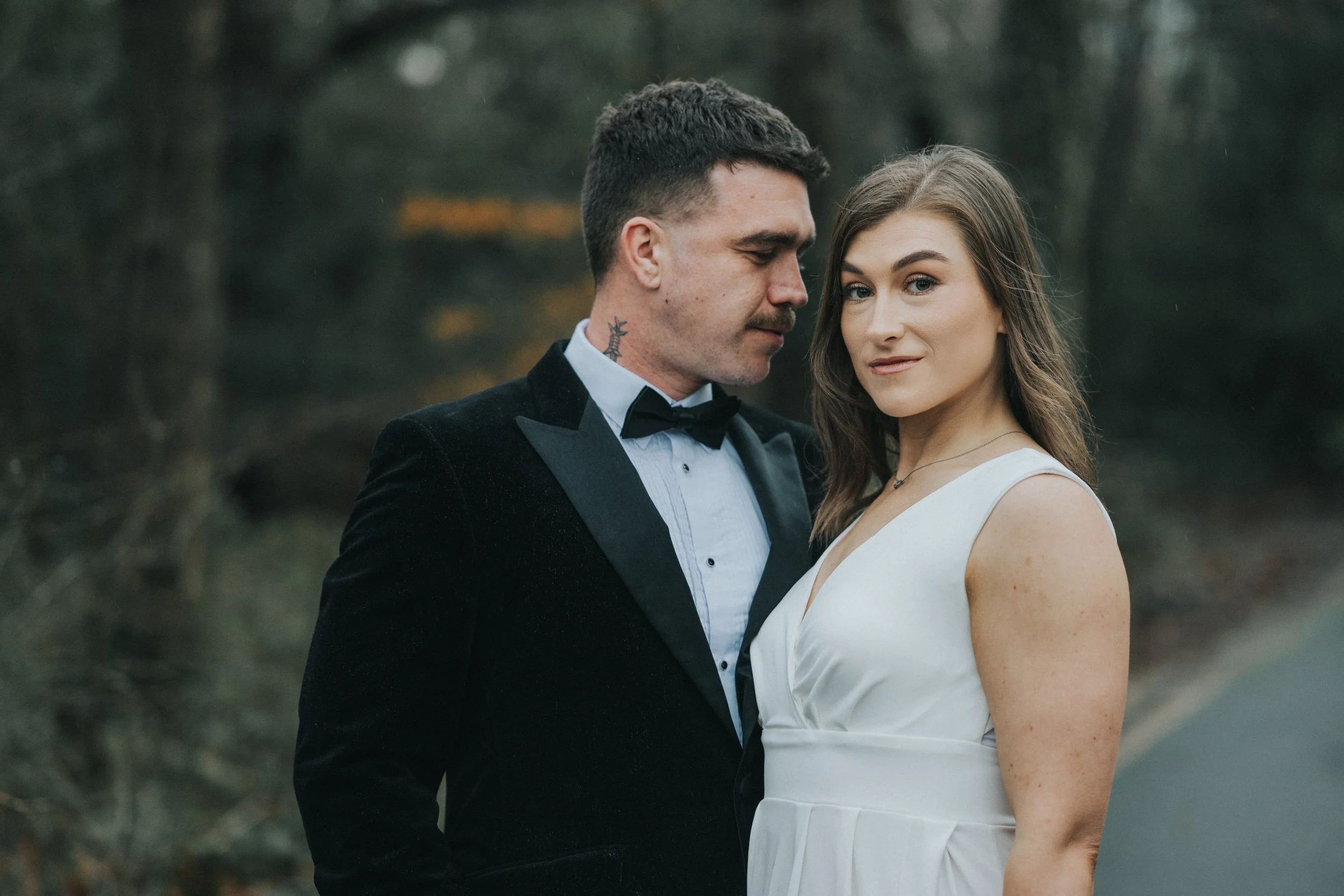 A man in a tuxedo and a woman in a white dress stand close together outdoors in a wooded area, looking at the camera.