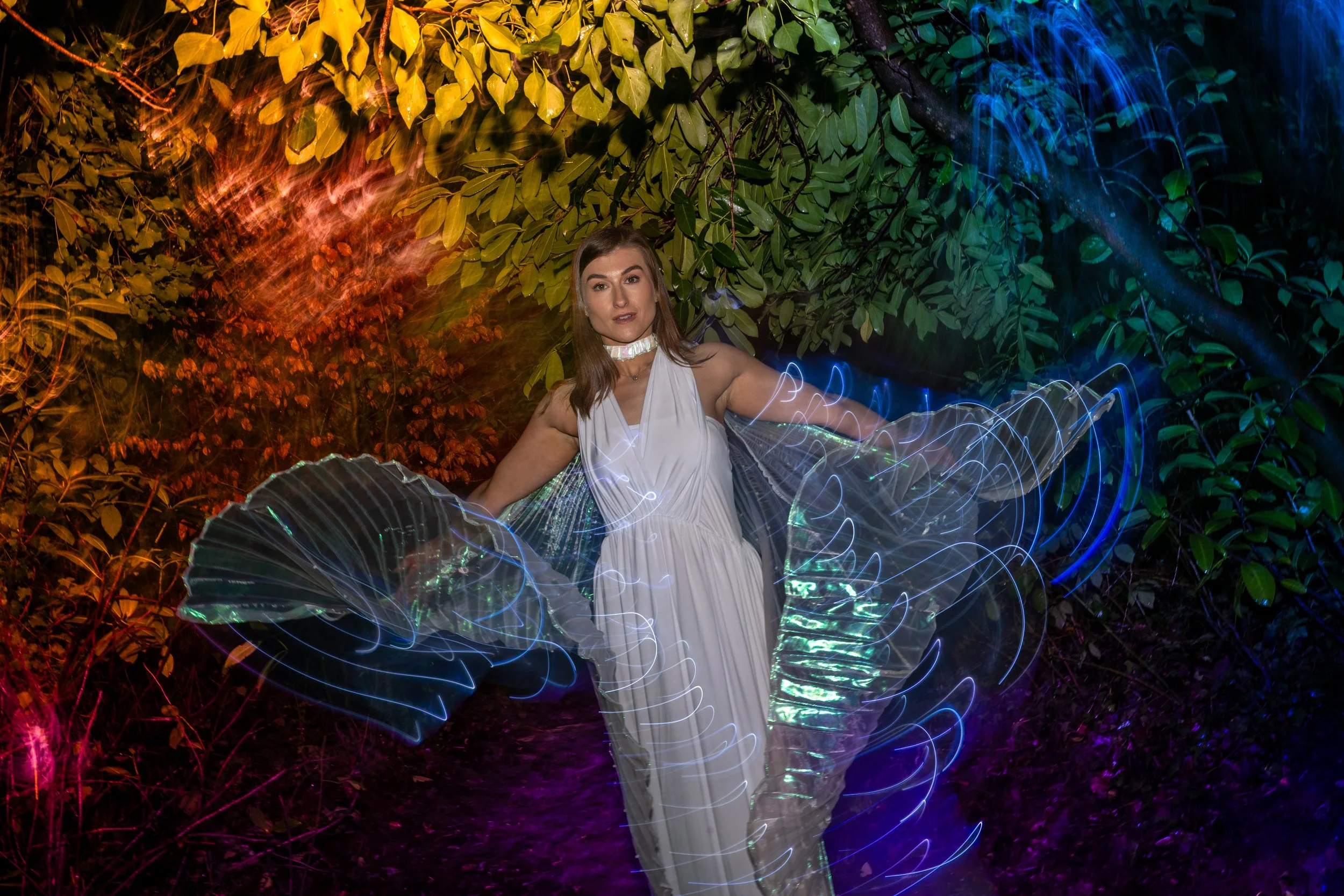 A woman in a white dress standing amidst colorful light trails and foliage, with dark night in the background.