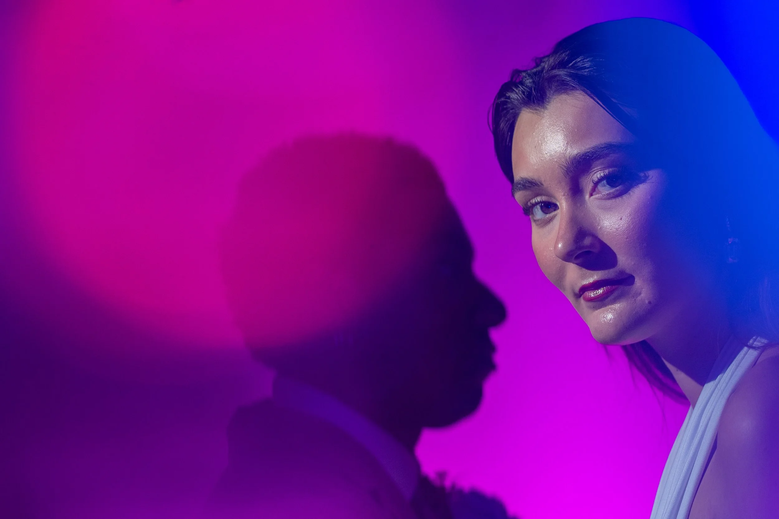 Half of a woman's face with short hair, highlighted by purple and blue lighting, looking at a shadowed silhouette of a person's face.