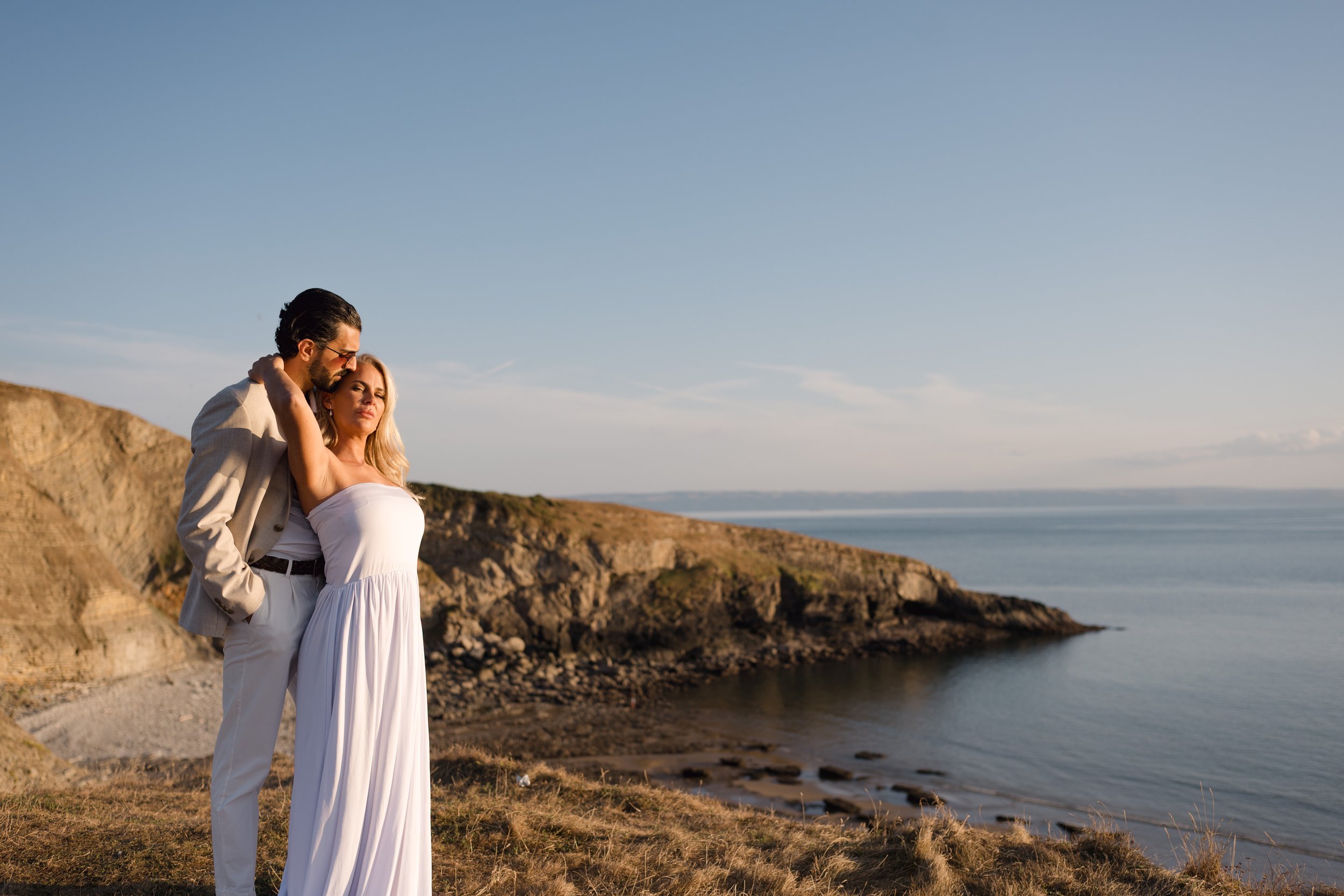 A couple stands on a rocky cliff near the ocean during sunset, with the man gently holding the woman who is wearing a white dress and the man in a beige suit with sunglasses.
