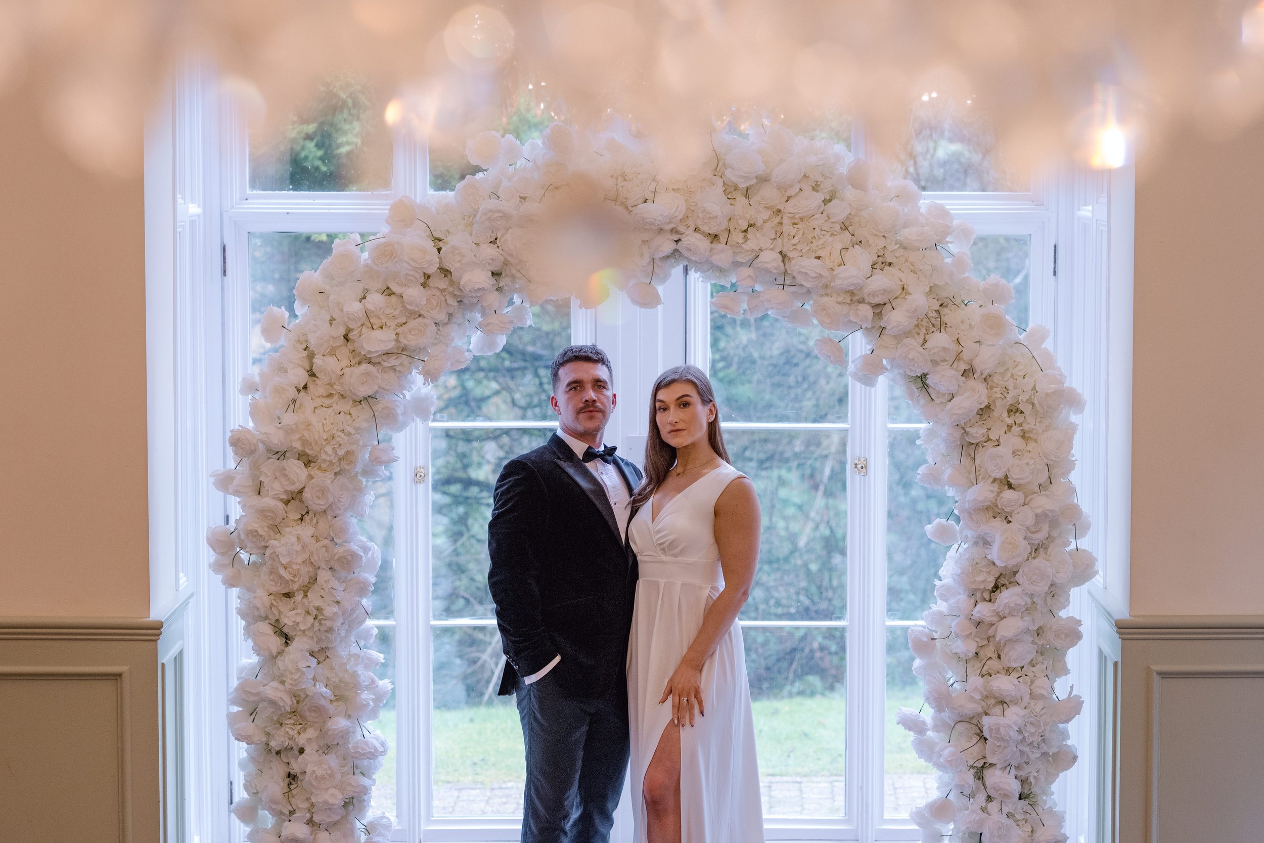 A couple in wedding attire standing under a large white floral arch in front of a window with trees outside.