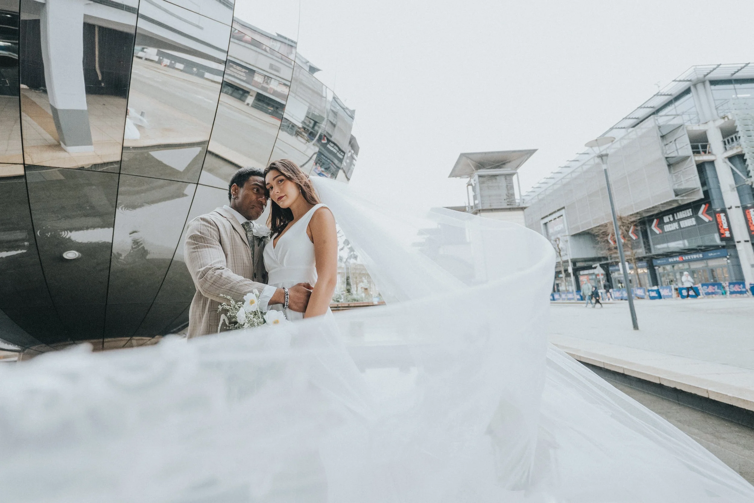 A couple dressed in wedding attire leaning against a reflective, modern building exterior with pedestrians and storefronts visible in the background.