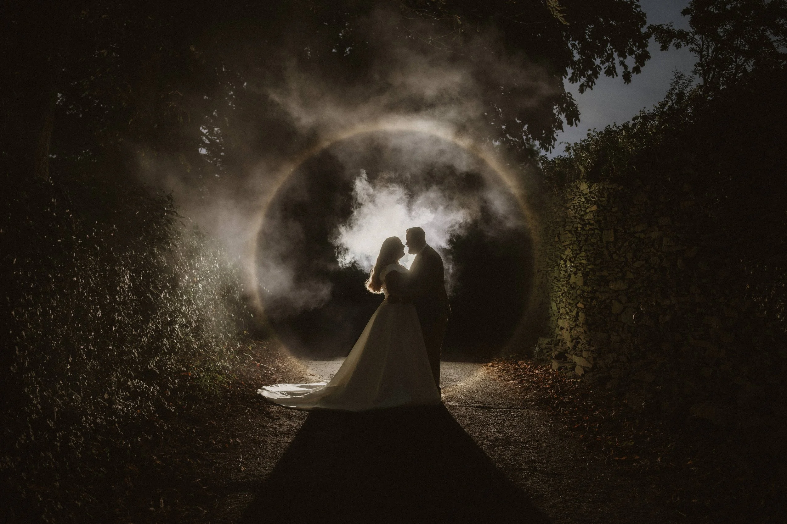 A silhouette of a couple in wedding attire standing close, holding each other, at night on a dirt path surrounded by trees and stone walls. They are backlit with a dramatic circle of light and mist behind them.