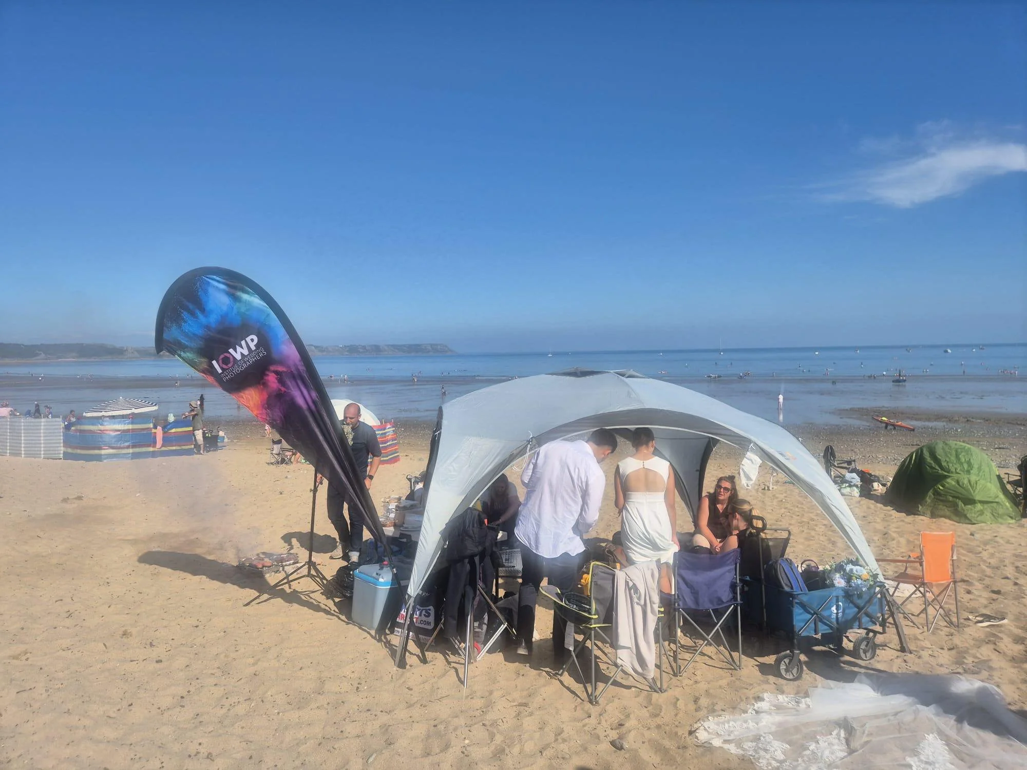 People gathered under a white canopy tent on a sandy beach with the ocean in the background. There are chairs, bags, and a green tent nearby, with some individuals standing and sitting, enjoying a day at the beach.