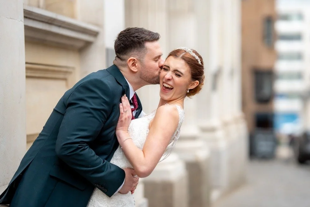 A happy bride and groom sharing a kiss with the groom kissing the bride on the cheek on a city street.