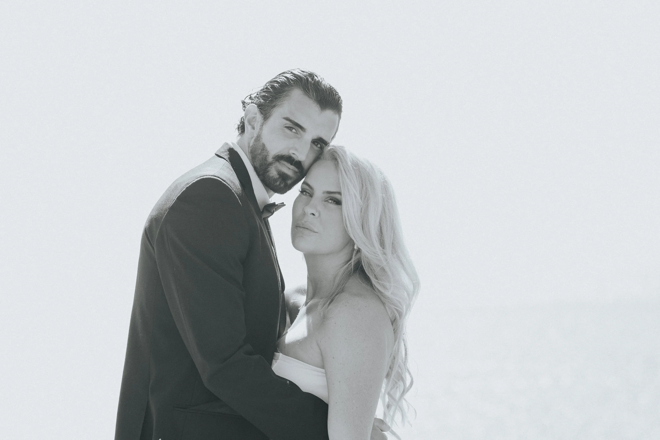 A black and white photograph of a man in a tuxedo and a woman in a strapless dress, embracing each other closely.