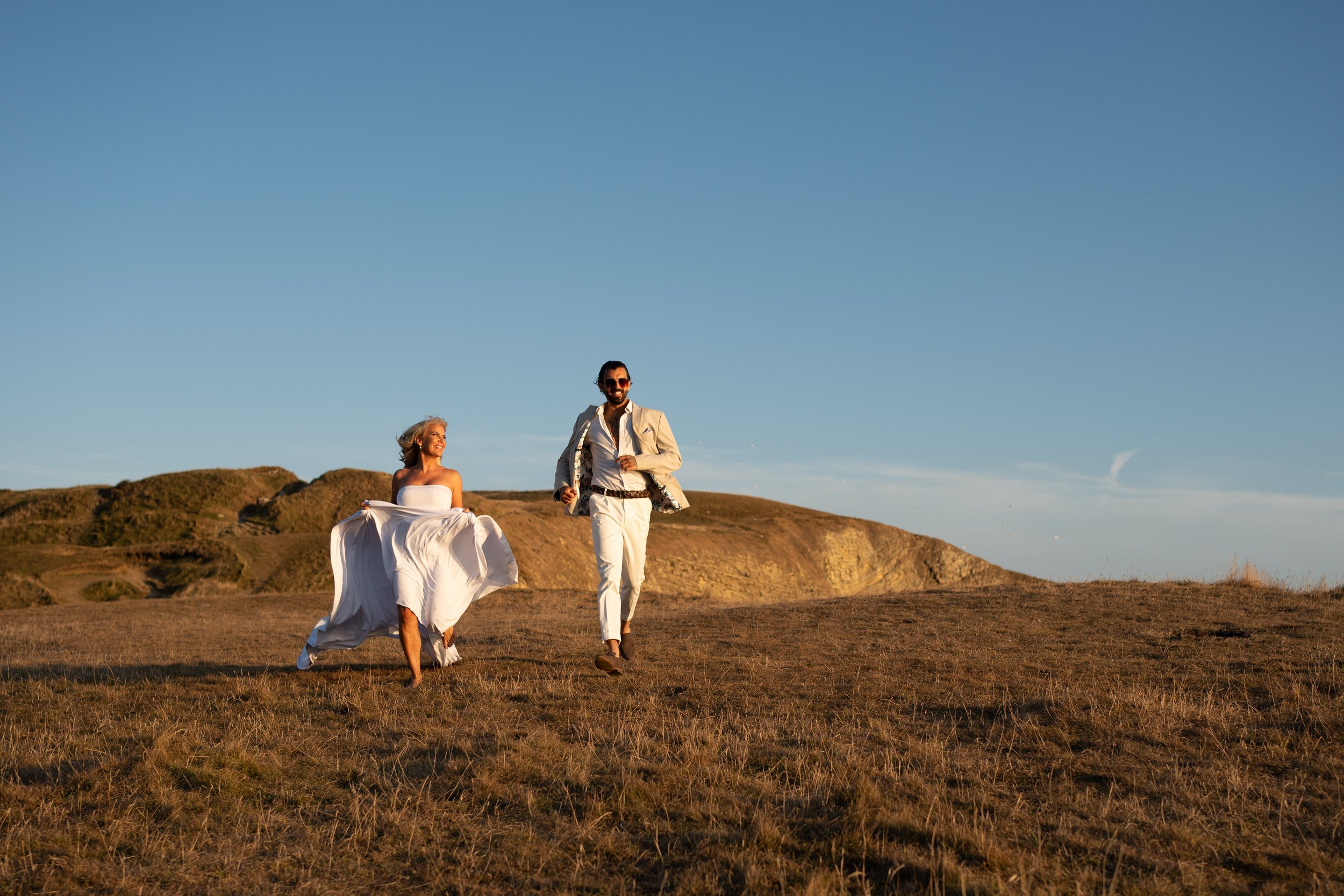 A woman in a white dress and a man in a white suit walk across a grassy hill during sunset, with clear blue sky and distant hills in the background.