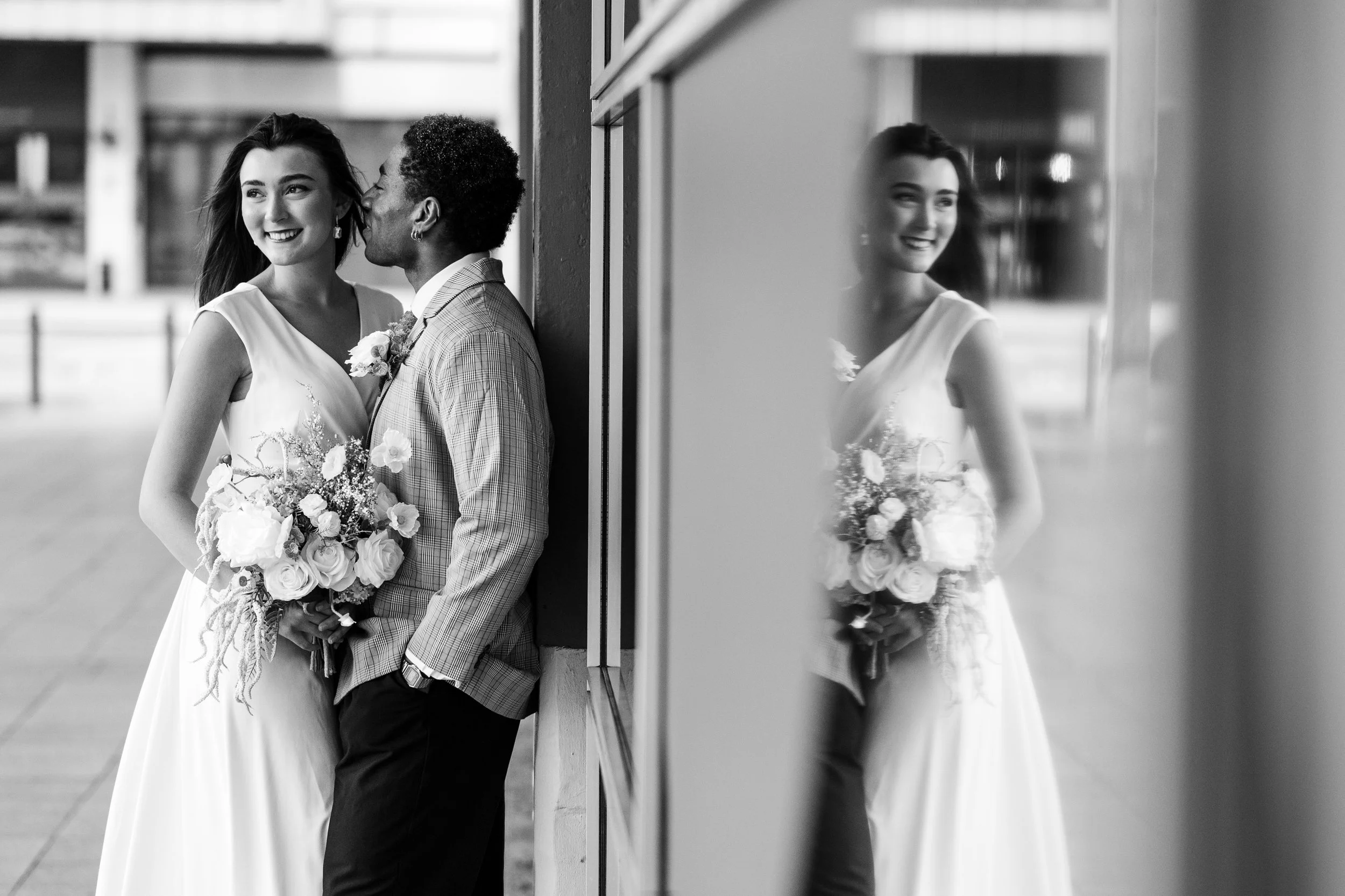 A black and white photo of a bride and groom leaning against a wall, smiling at each other, with the bride holding a bouquet. The reflection of the bride is visible on a glass surface to the right.