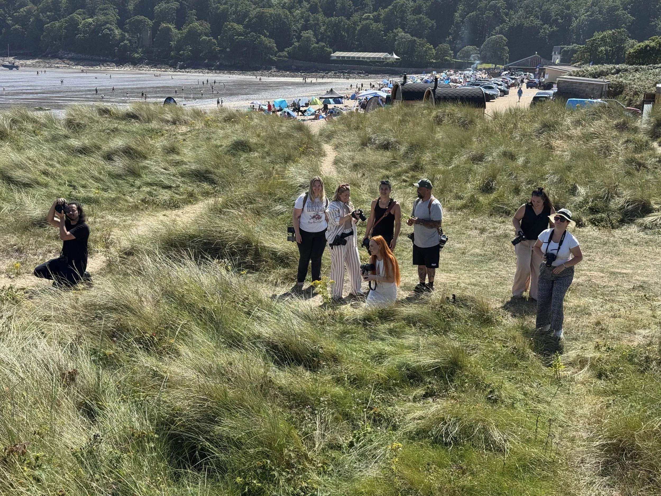Group of people with cameras standing and sitting on grassy dunes near a sandy beach and ocean in the background.