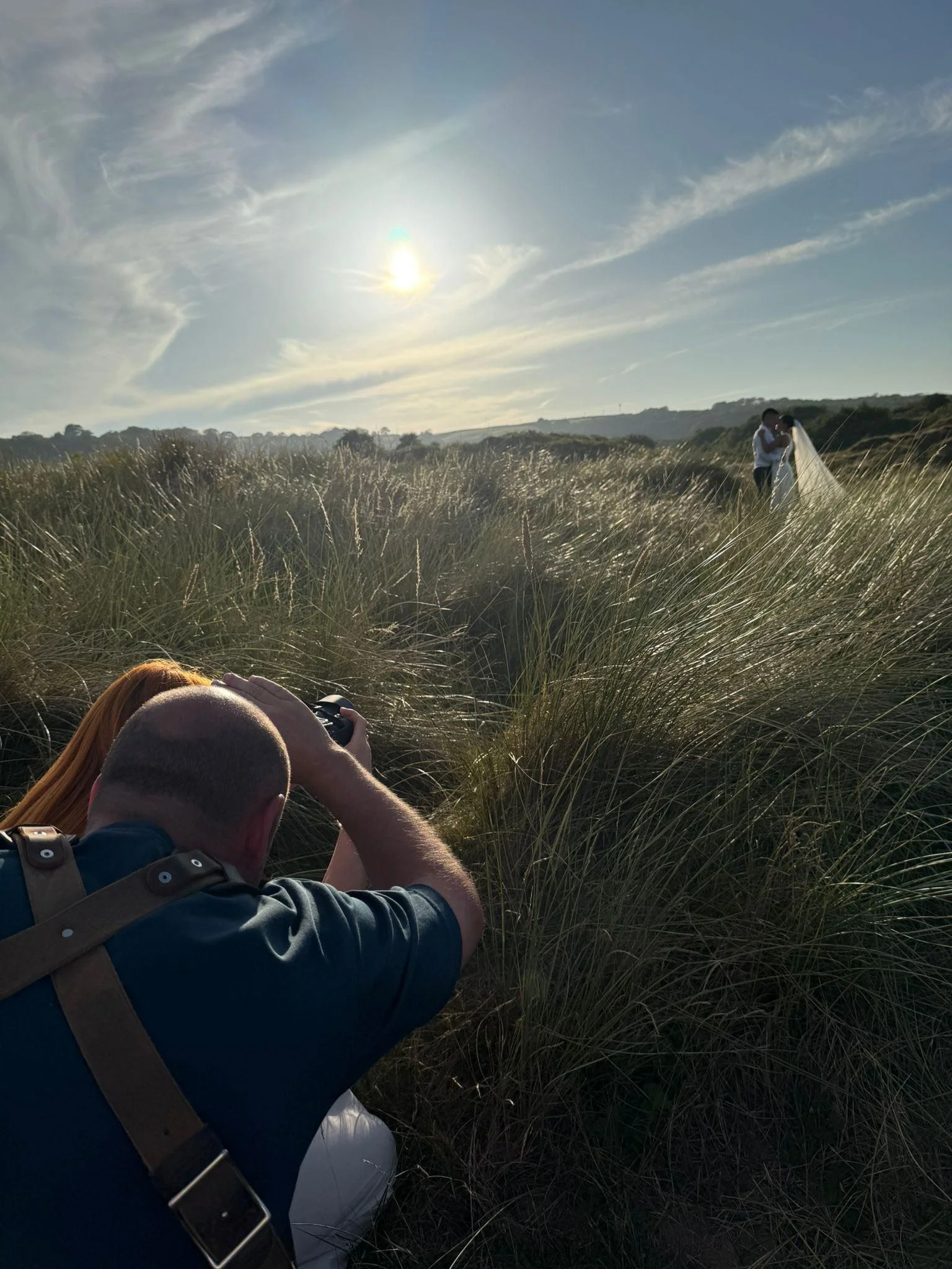 A couple is standing closely together in a grassy field, holding hands and facing each other, likely during a wedding or romantic photoshoot. The sun is shining brightly in the sky behind them, casting a warm glow.