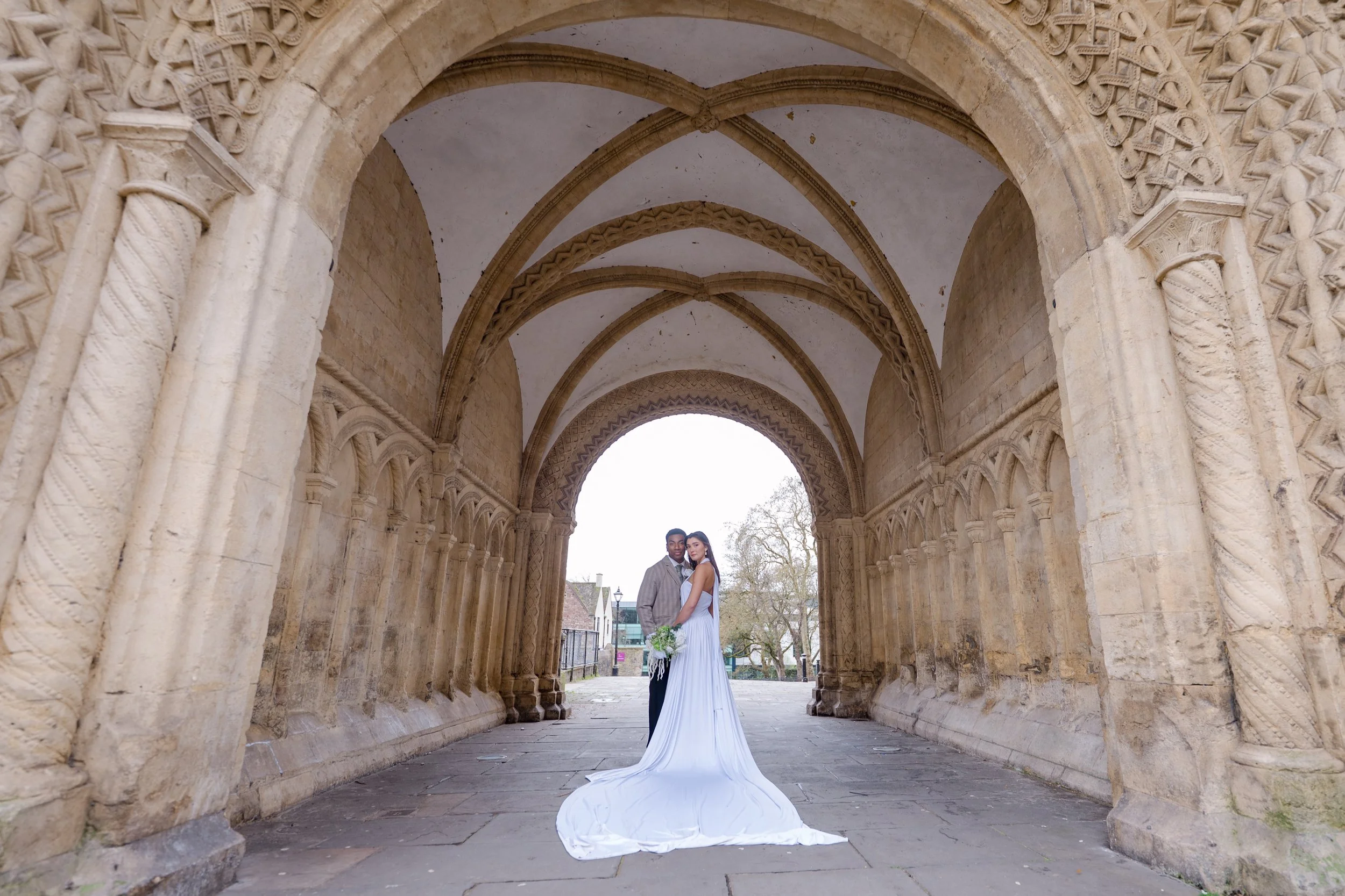 A bride and groom standing together under an ornate stone archway at a historic church or cathedral. The bride is wearing a white wedding gown and holding a bouquet, and the groom is in a gray suit. The background shows trees and a street scene.