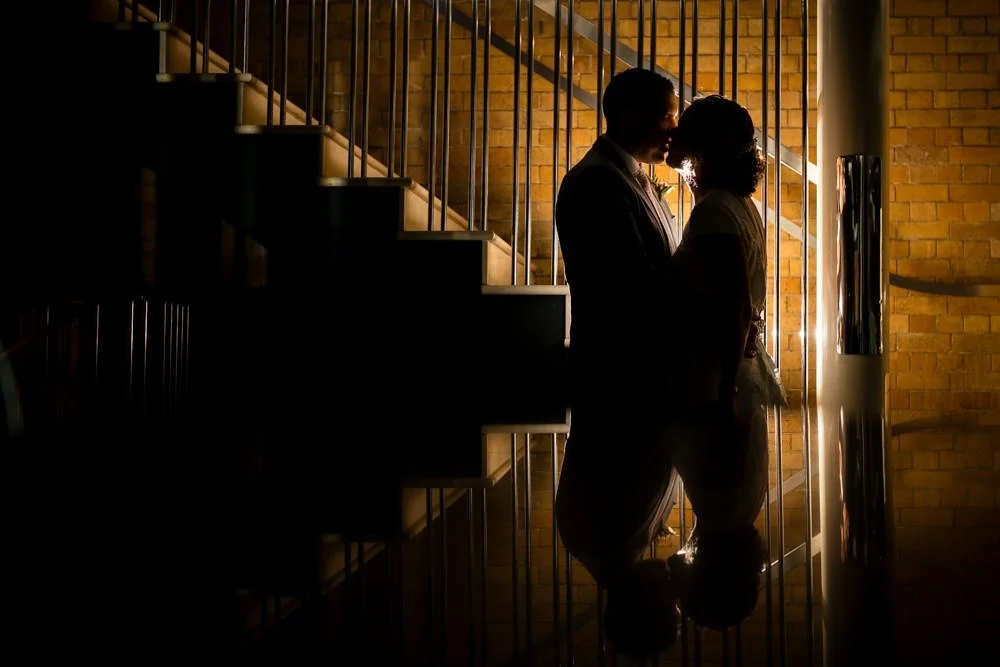 Silhouette of a couple standing close together, facing each other, in front of a brick wall and staircase, backlit with warm lighting.