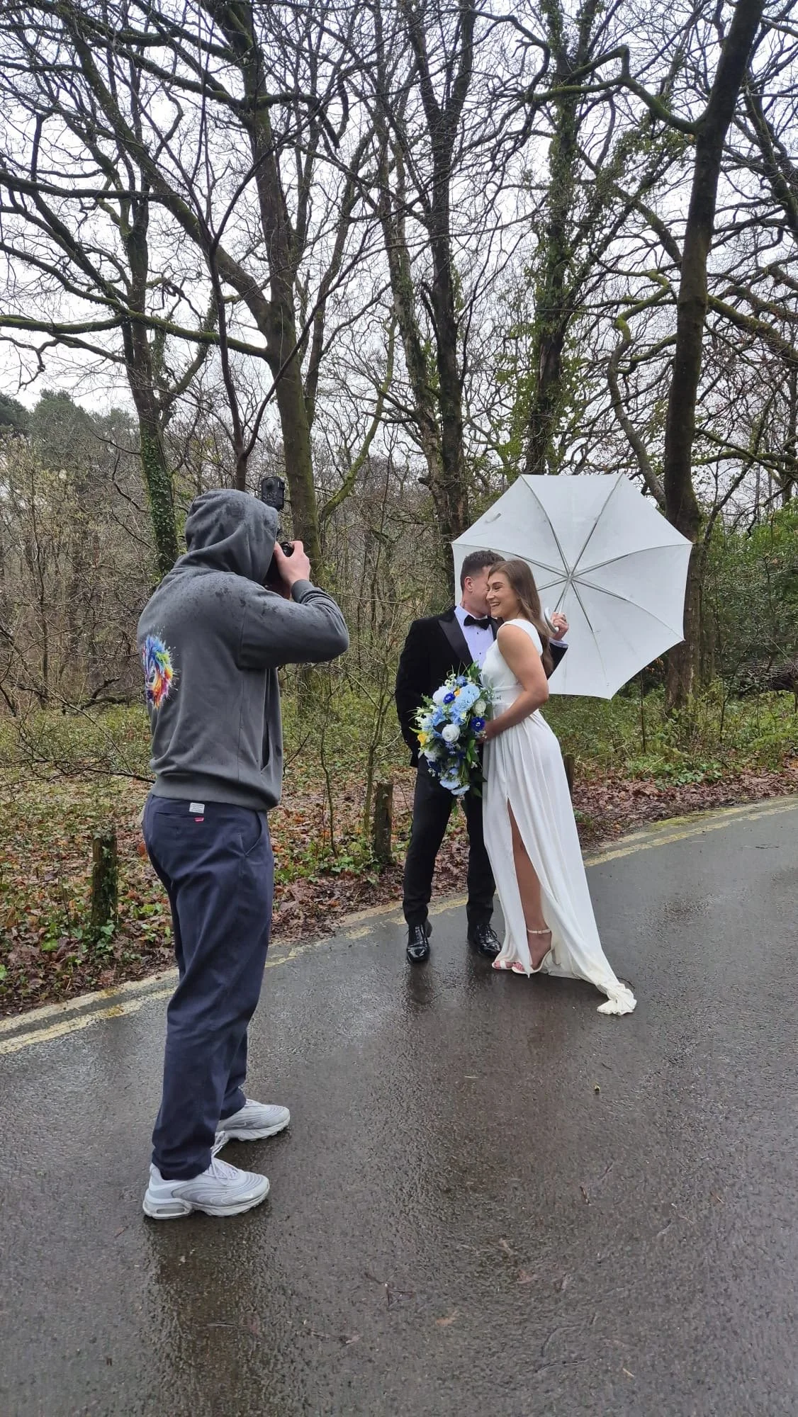 A bride and groom in wedding attire sharing a kiss outdoors, with a photographer taking their picture and holding a white umbrella on a rainy day in a wooded area.