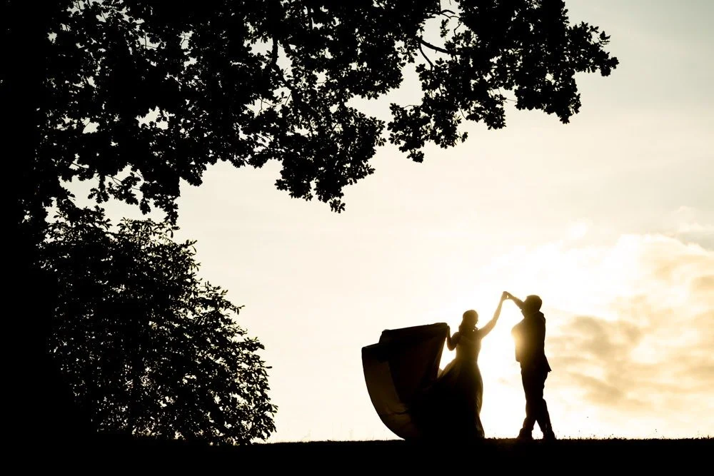 Silhouette of a couple dancing outdoors during sunset, with large trees in the background.