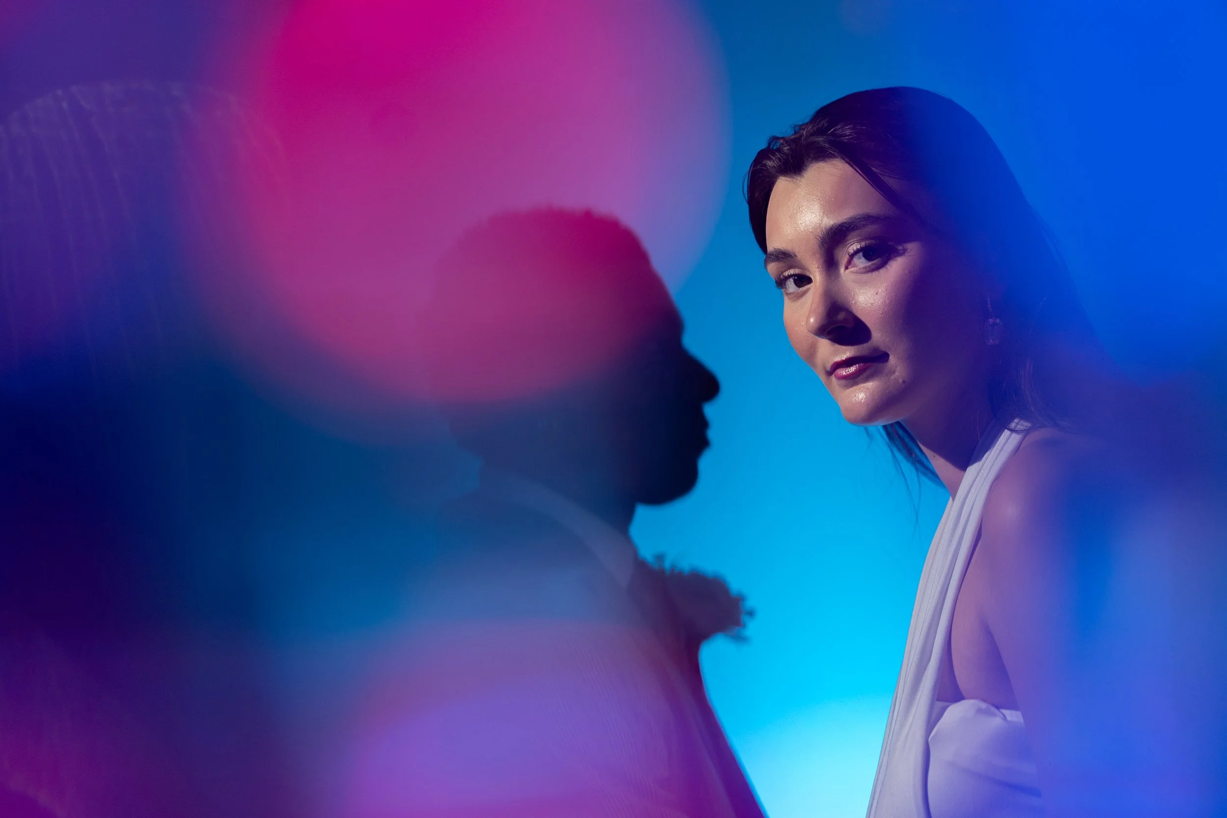 A woman with dark hair and earrings looks confidently at the camera with colorful, diffused pink and blue lighting around her.