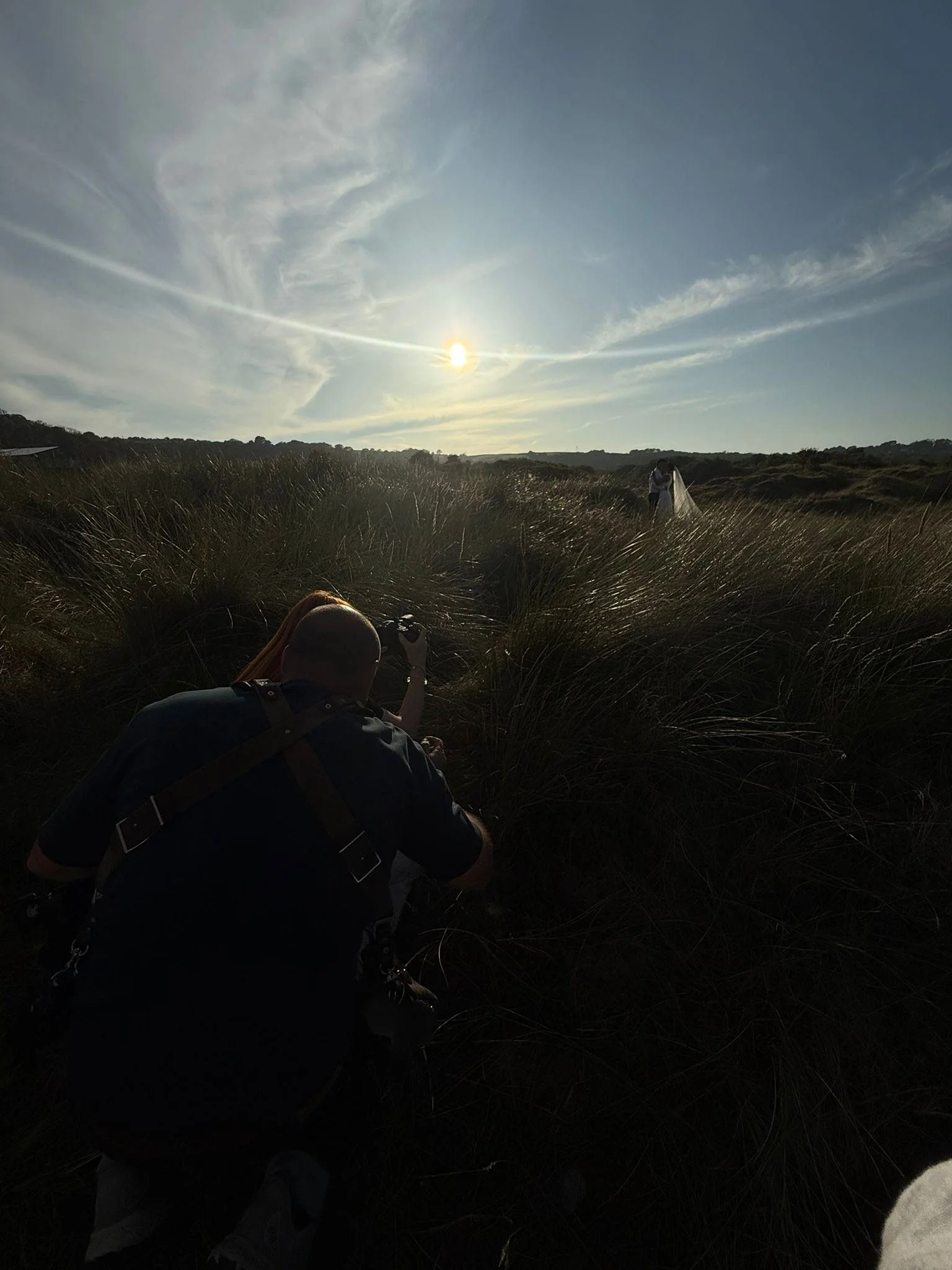 A person kneeling in tall grass taking a photograph of a bride in a white wedding dress and veil with the sun setting in the background.