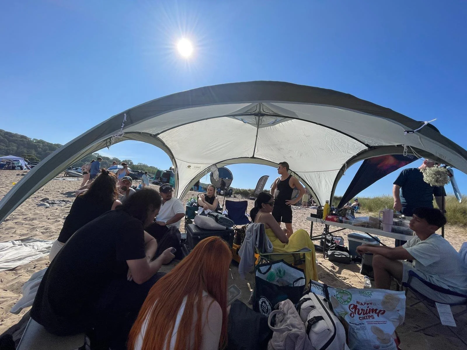 Group of people gathered under a beach tent on a sunny day, with a clear blue sky, in a sandy outdoor area.