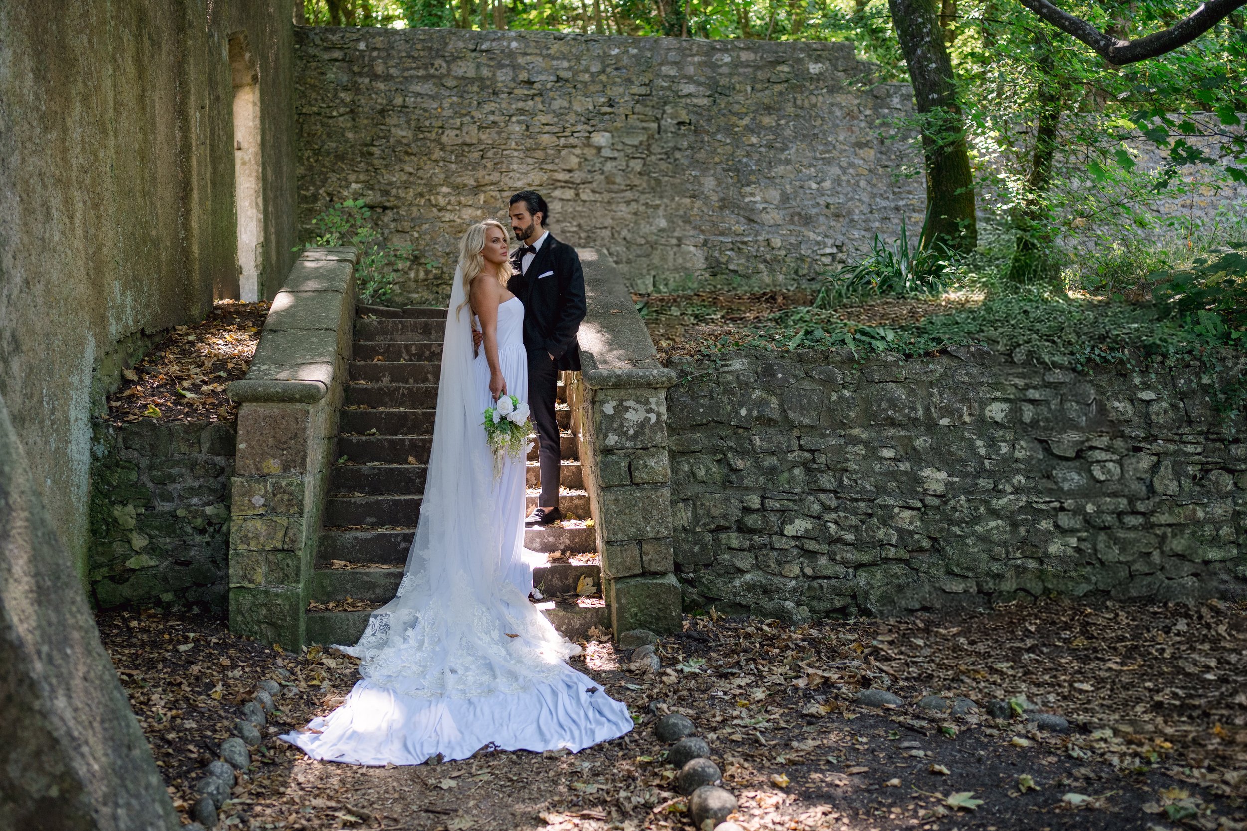 A bride and groom standing on stone steps in a wooded outdoor setting, with trees and a stone wall in the background. The bride is in a white wedding dress holding a bouquet, and the groom is in a tuxedo. Sunlight filters through the trees, illuminat
