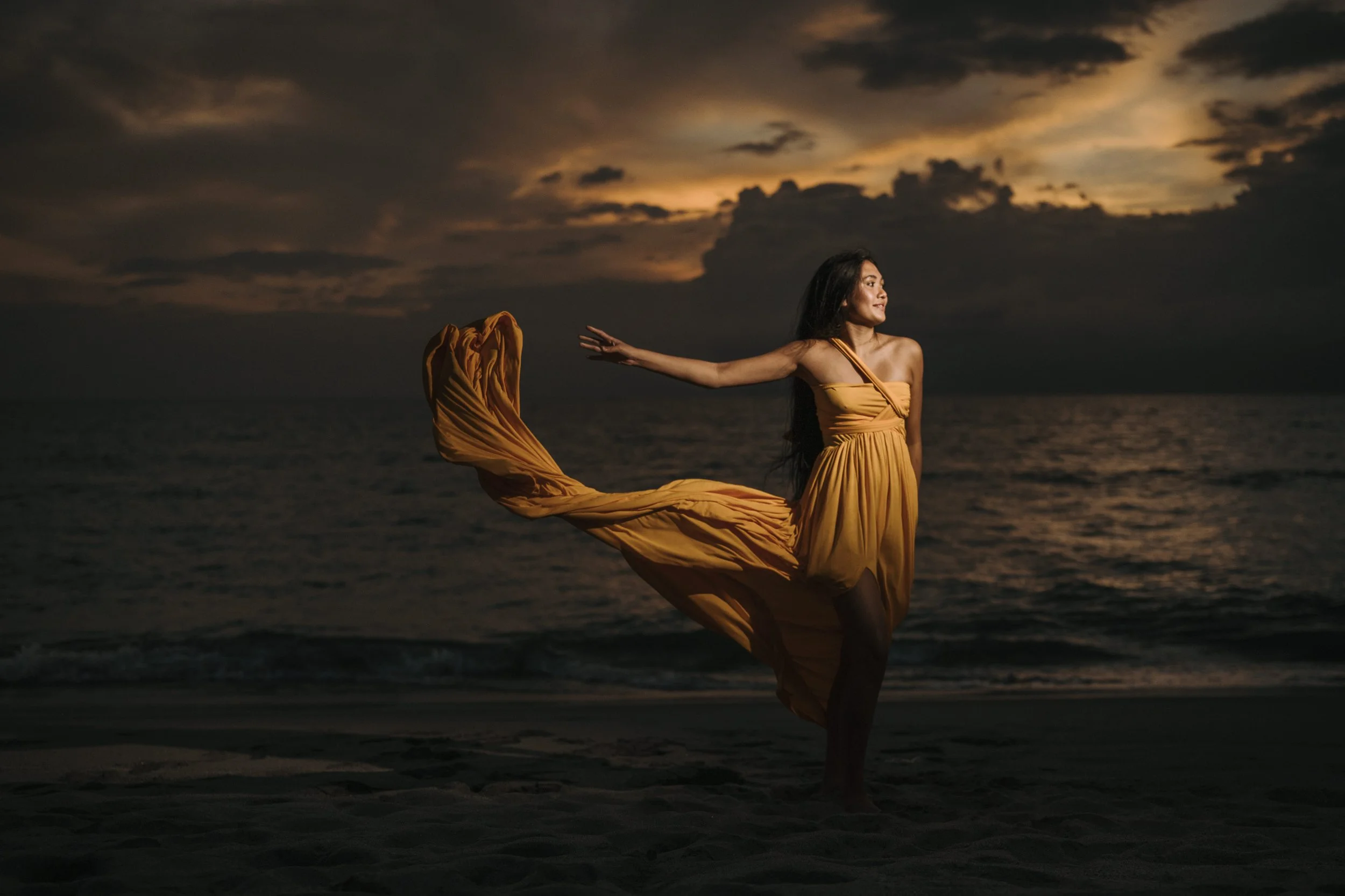 Woman in a flowing yellow dress performing a dance on the beach at sunset with a dramatic cloudy sky in the background.