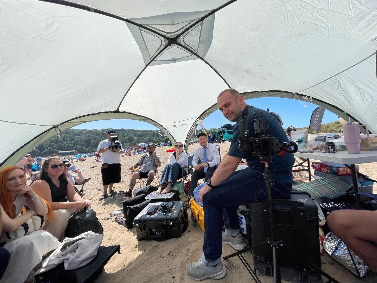 People sitting inside a beach tent with cameras and equipment, on a sandy beach with many beachgoers and hills in the background.