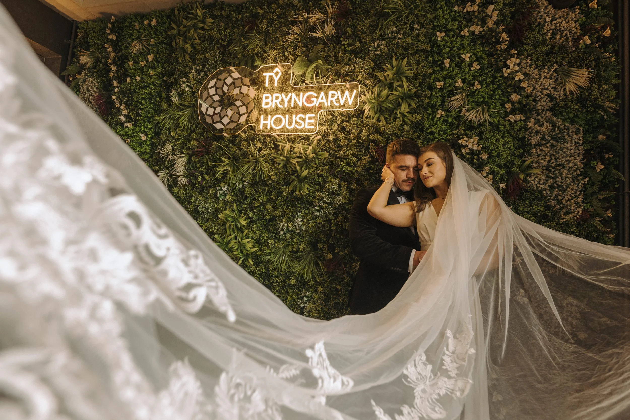 A bride and groom embracing underneath a lush green wall with a neon sign that reads 'TY BRYNGARW HOUSE' in cursive and block letters, with the bride wearing a long veil and the groom in a black suit.