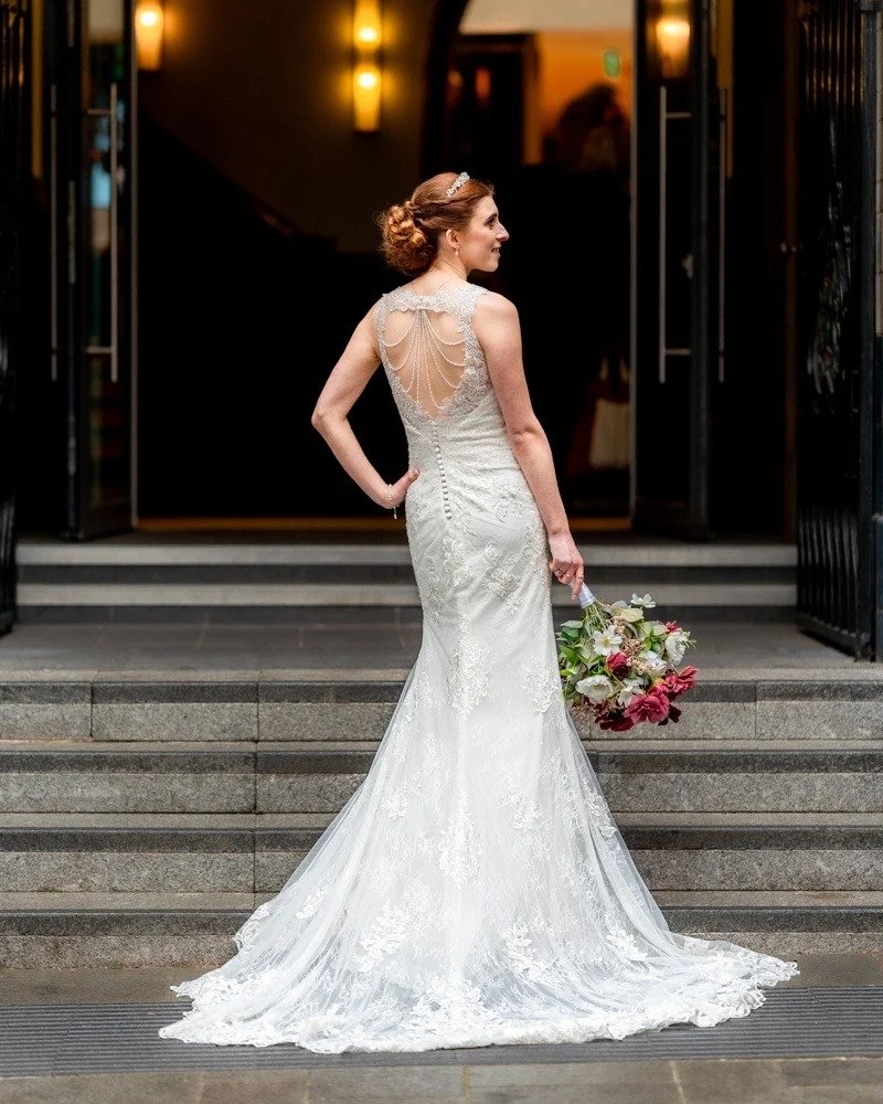 Bride in a white lace wedding gown holding a bouquet of flowers, standing on stairs outside a building at night.