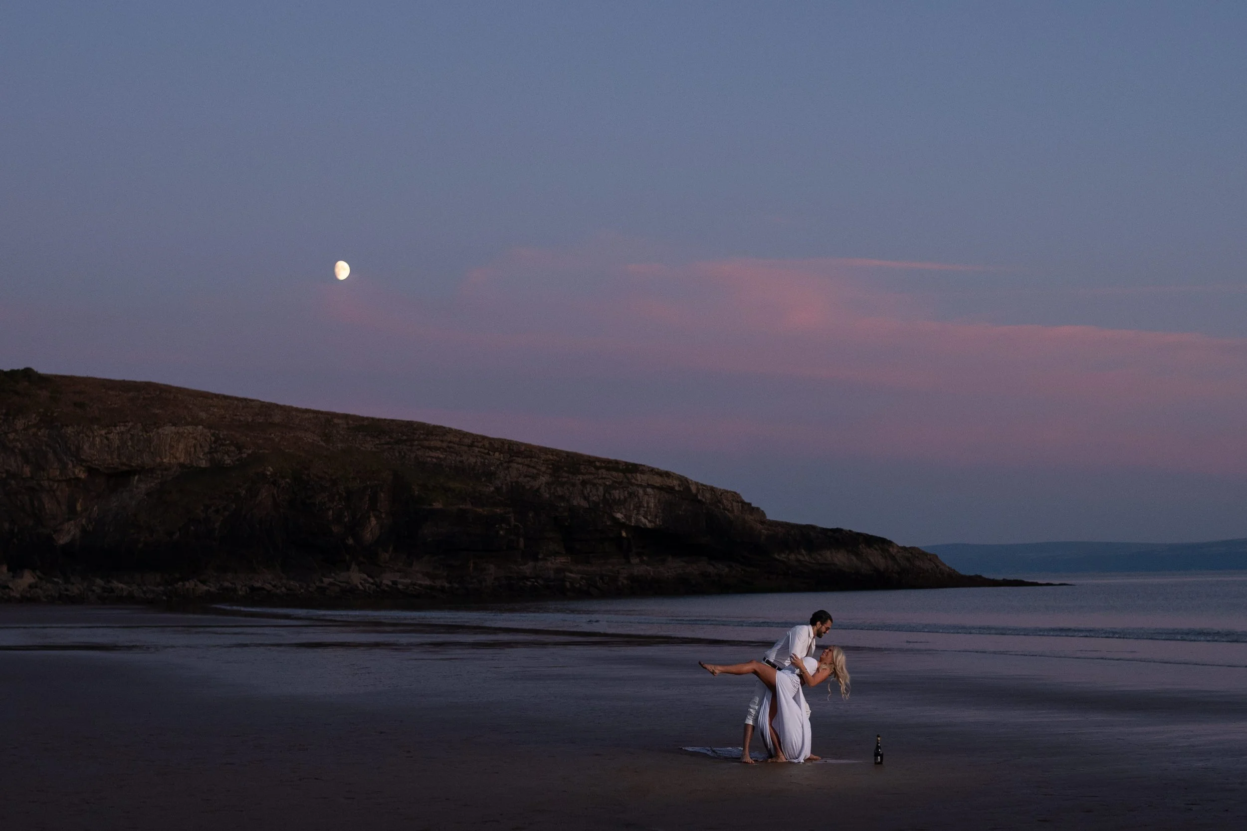 A couple dressed in white, with the woman lying on her back and the man holding her, on a beach at sunset with a full moon in the sky and mountains in the background.