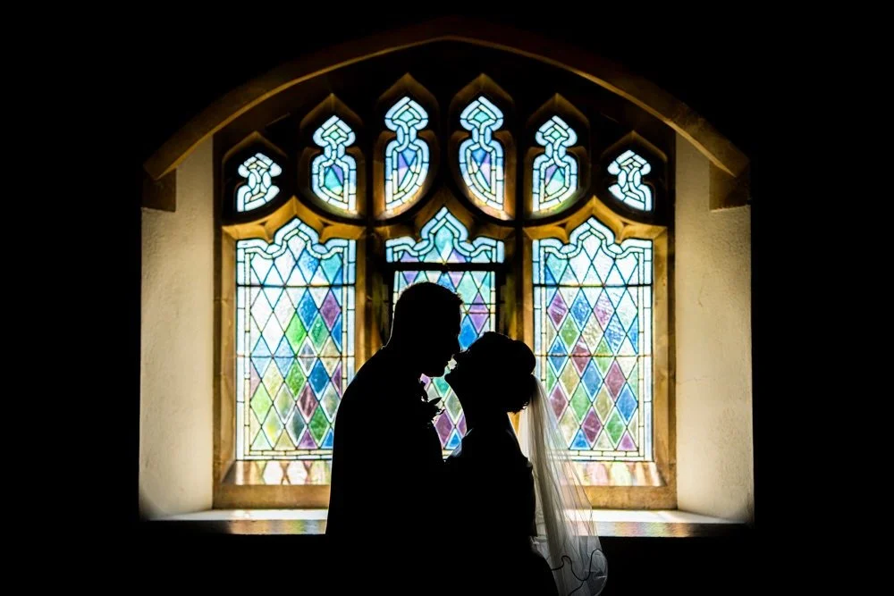 Silhouette of a bride and groom standing close together in front of a colorful stained glass window.