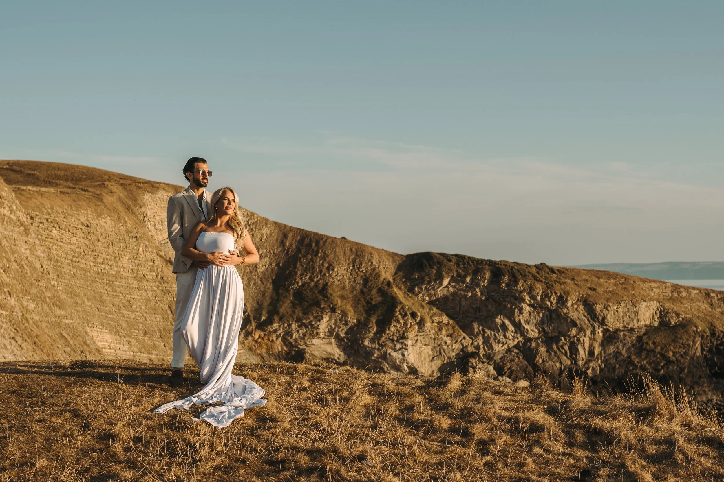 A couple dressed in formal attire standing on a grassy cliff overlooking the ocean, with rugged cliffs in the background, during sunset.