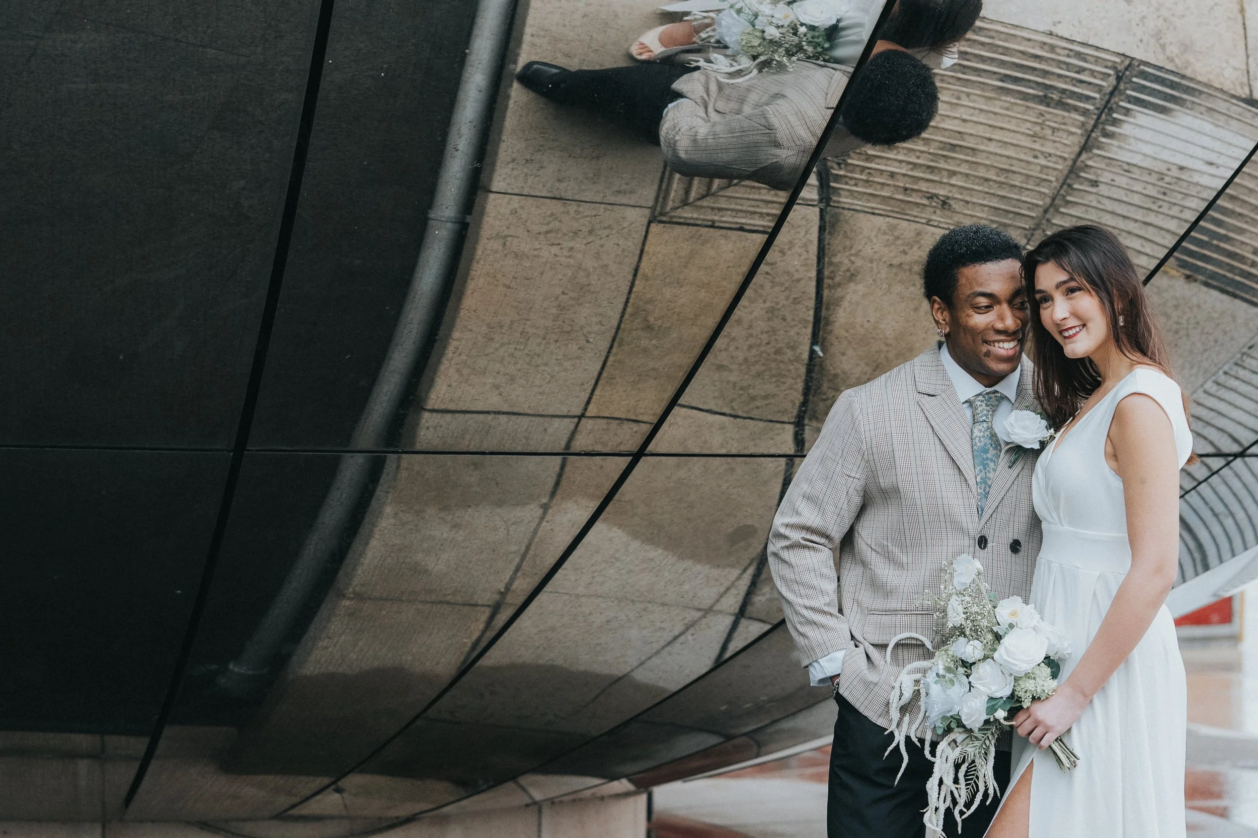 A newlywed couple standing close together and smiling, the groom dressed in a gray checkered suit with a white flower boutonniere and the bride in a sleeveless white dress, holding a bouquet of white flowers, standing outside near a building with ref