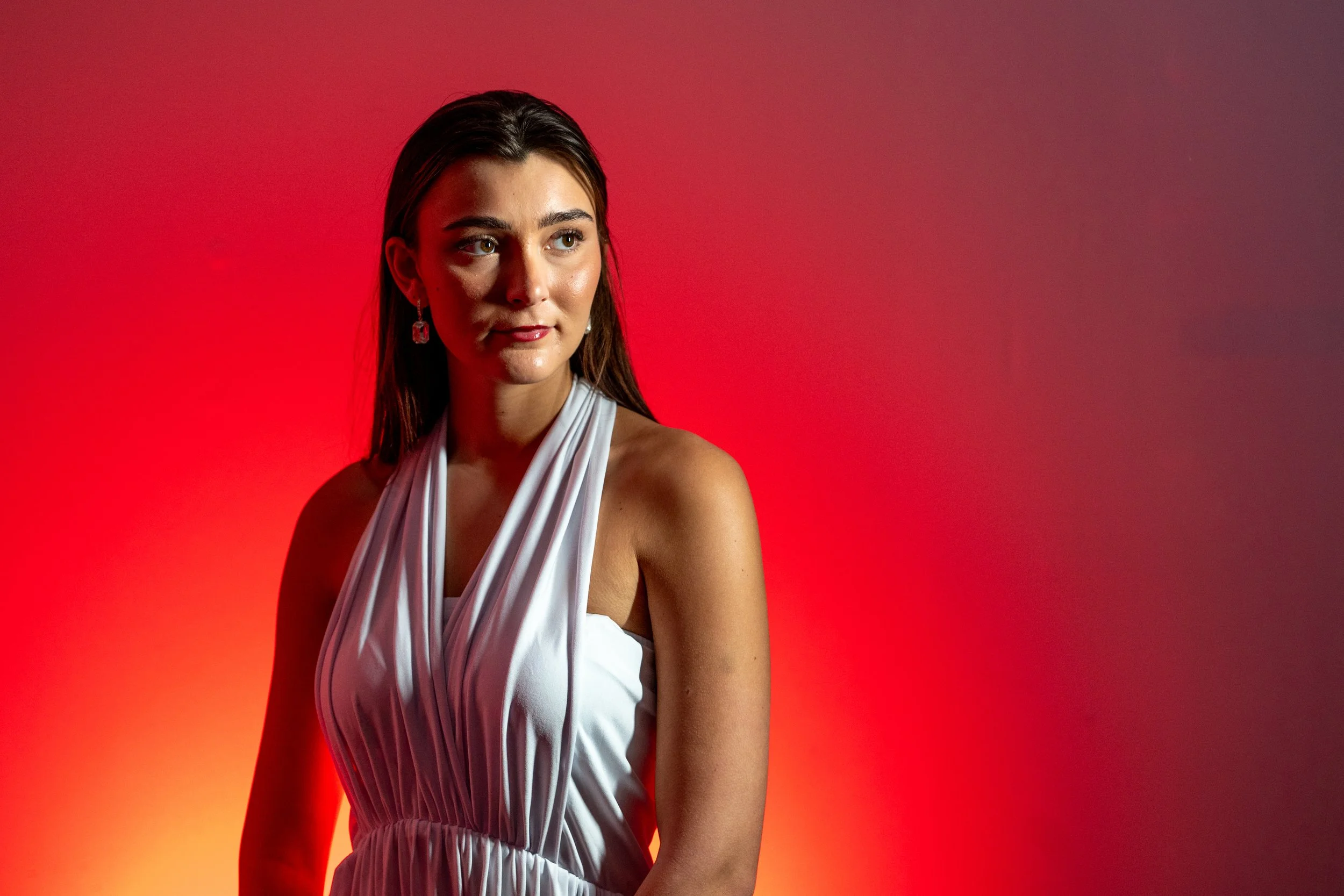 A woman with dark hair and tan skin wearing a white dress with draped fabric, standing against a red and orange gradient background.