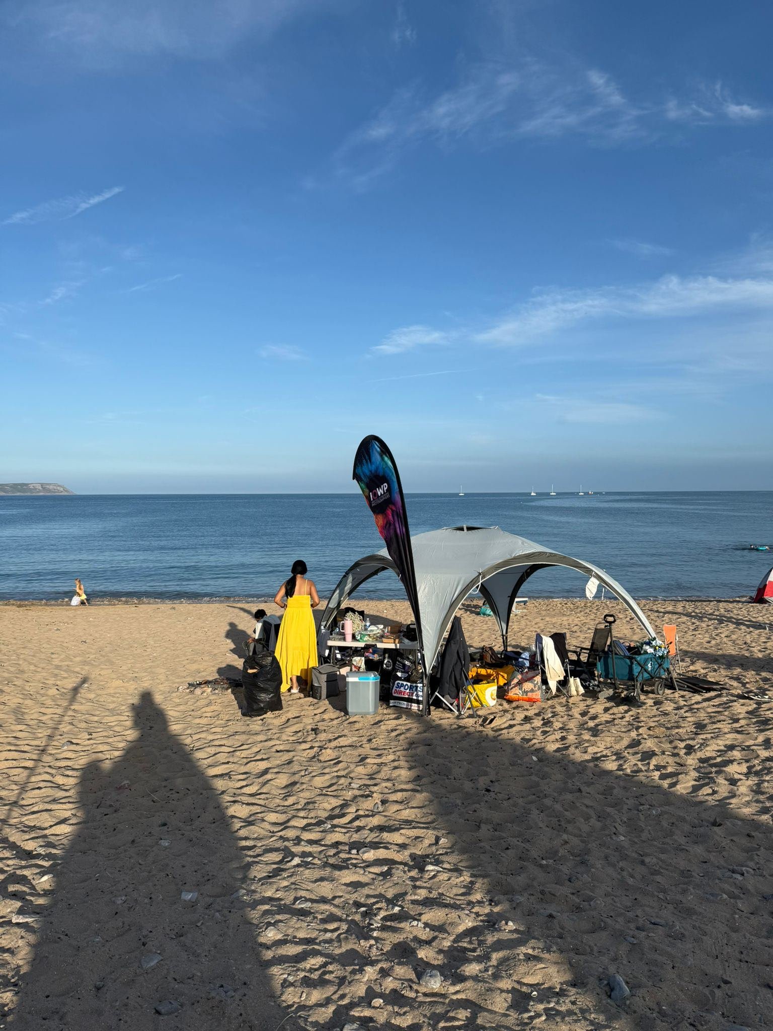 Beach scene with a group of people under a tent, some sitting and some standing, with sand, ocean, and blue sky in the background.