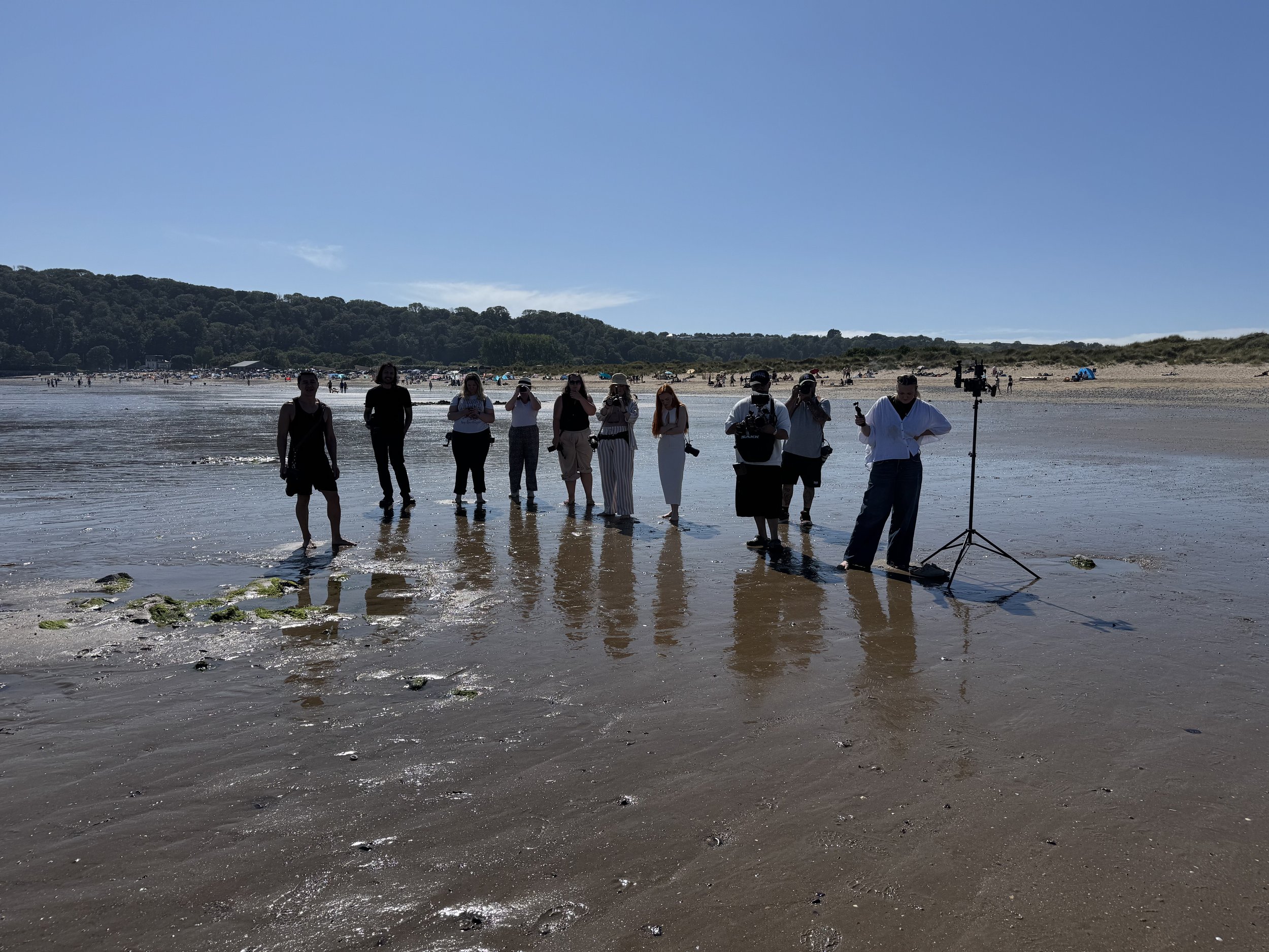 Group of people on a beach, some holding cameras, near a tripod with equipment, during daytime with a clear sky.