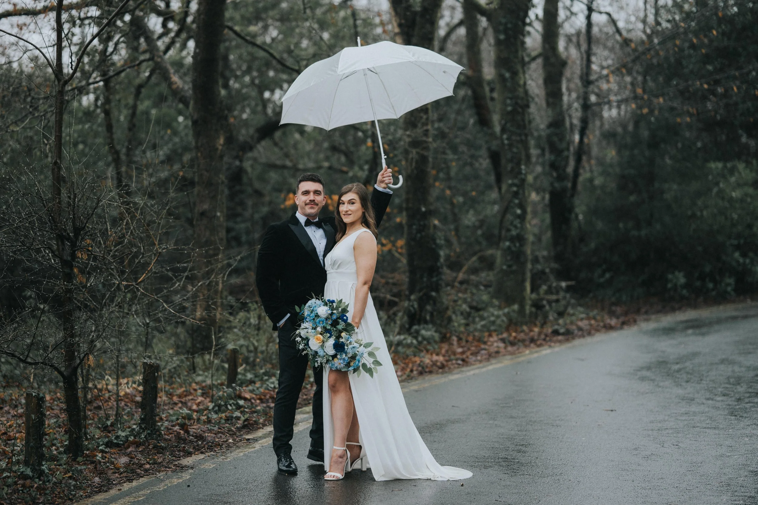 Wedding couple standing on wet road in a wooded area, with the bride holding a bouquet of blue and white flowers and the groom wearing a tuxedo, while the bride holds a white umbrella over them.