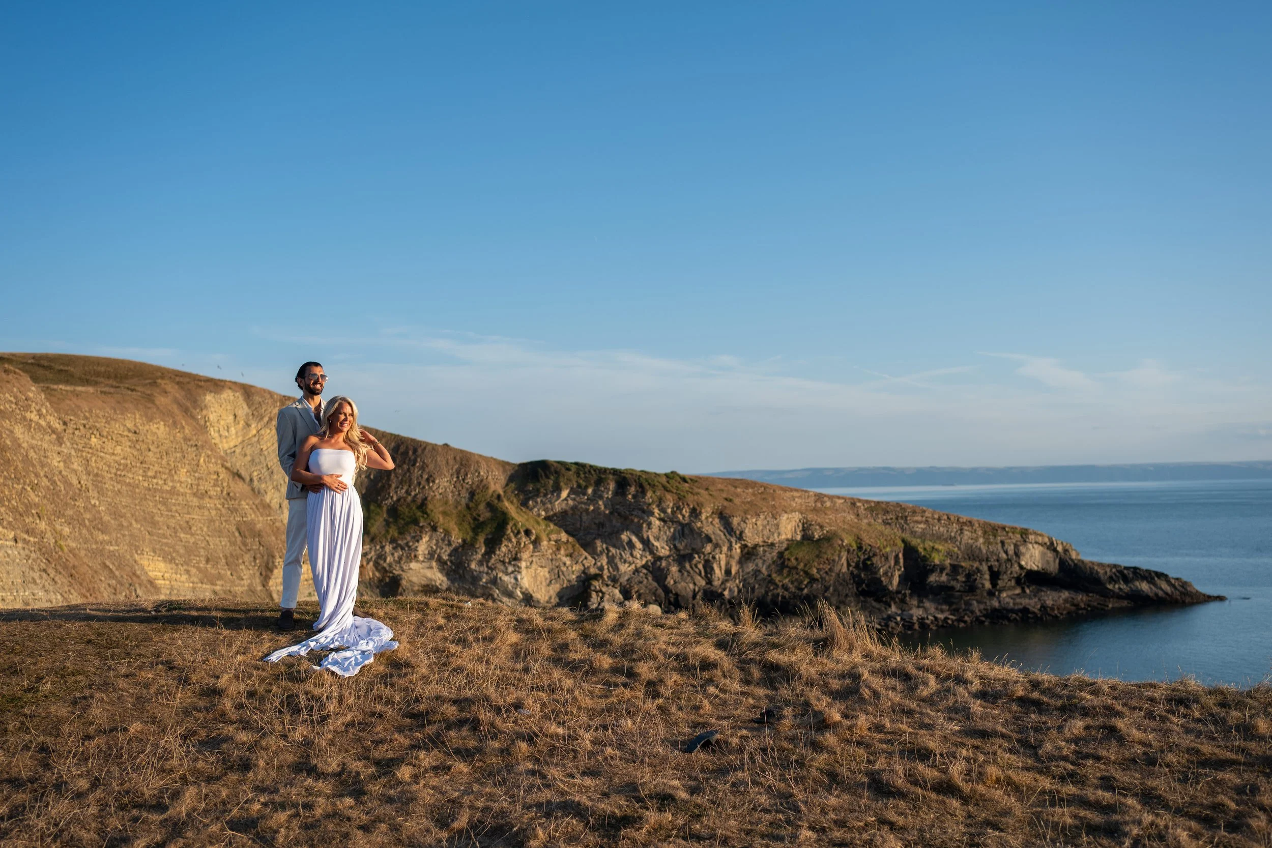 A couple, dressed formally, standing on a grassy cliff overlooking the ocean under a clear blue sky, with the woman wearing a white strapless dress and the man in a light-colored suit.