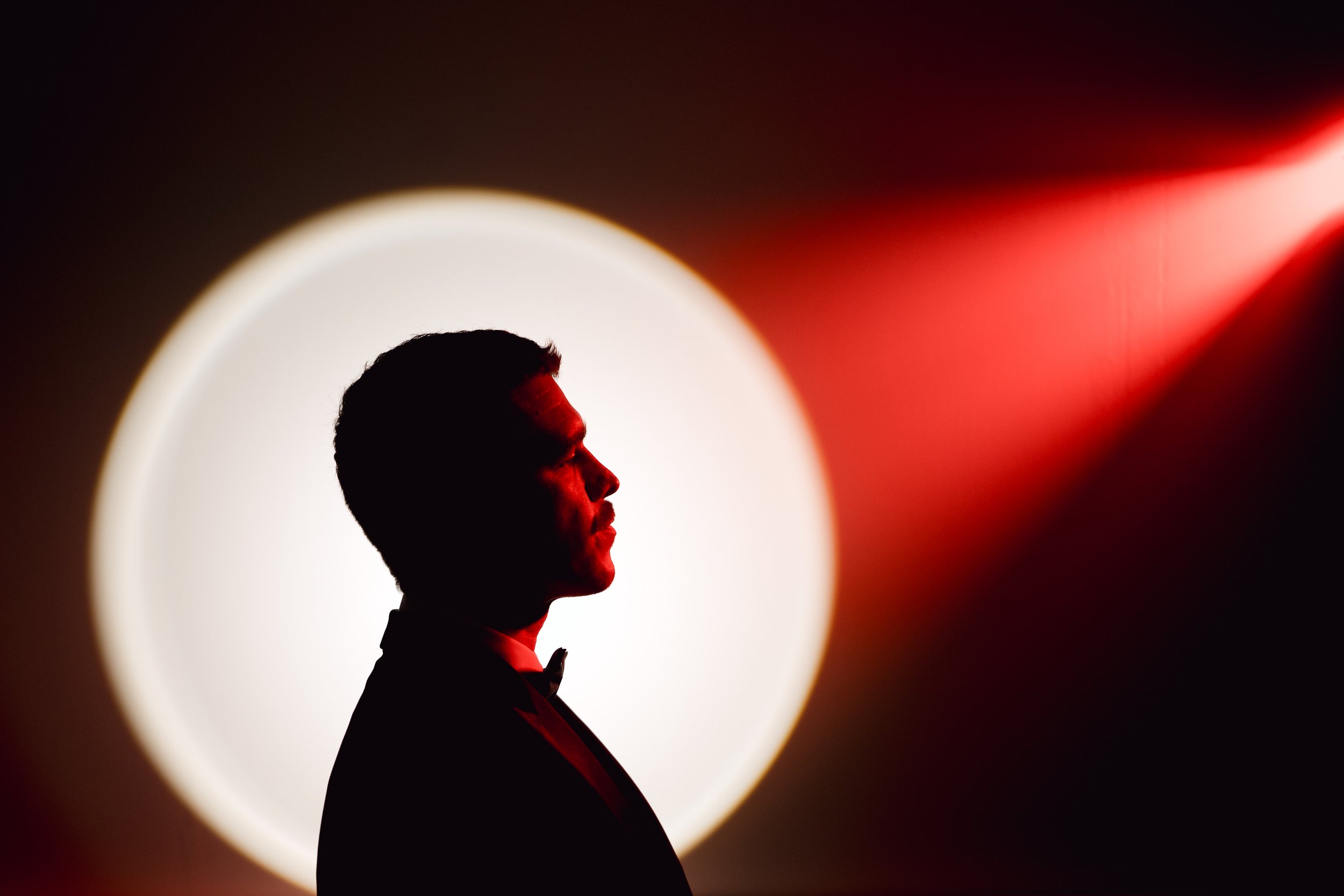Silhouette of a man in a tuxedo with a bow tie, standing in front of a large circular white light with red accents.