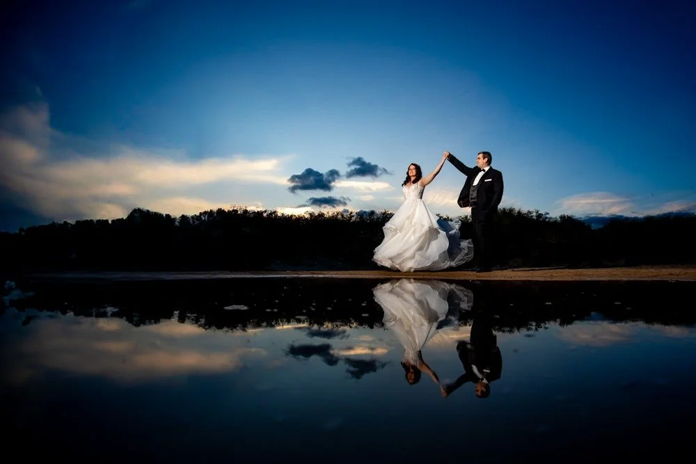 Bride and groom in wedding attire holding hands on a beach at sunset, with their reflection visible in the water.
