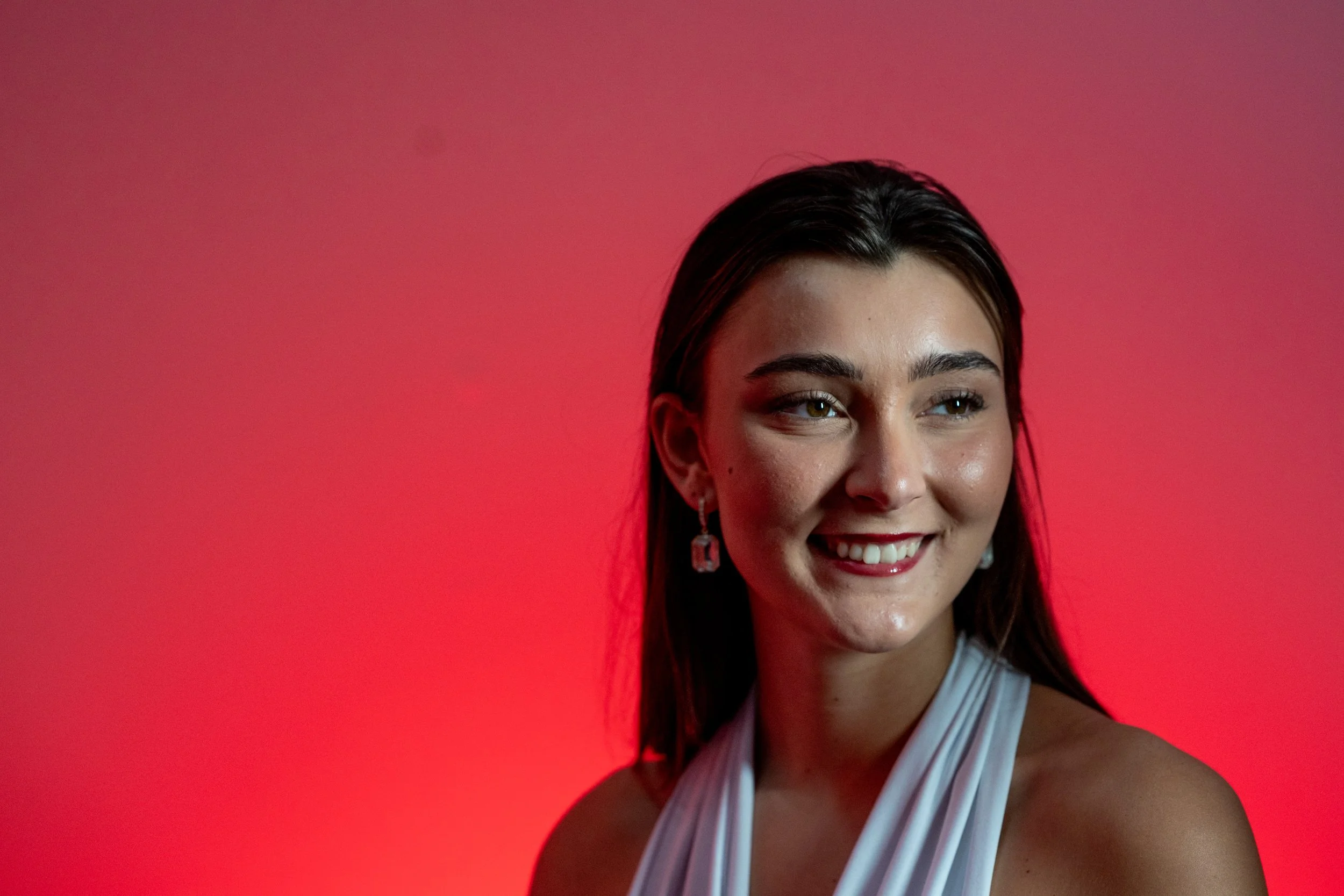 Close-up of a young woman with dark hair, smiling, wearing earrings, with a red background.
